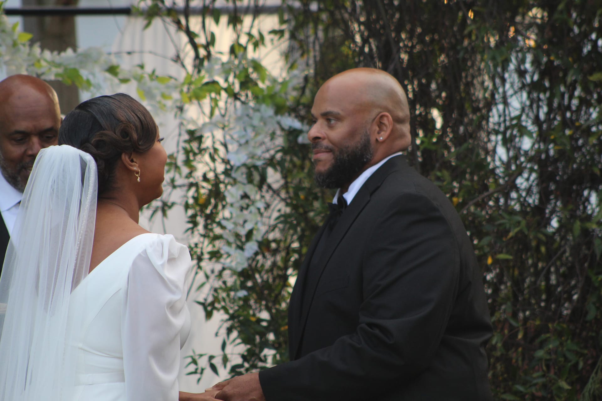 Groom in a black tuxedo smiles warmly at his bride during an outdoor wedding ceremony surrounded by lush greenery at Scarborough House estate.