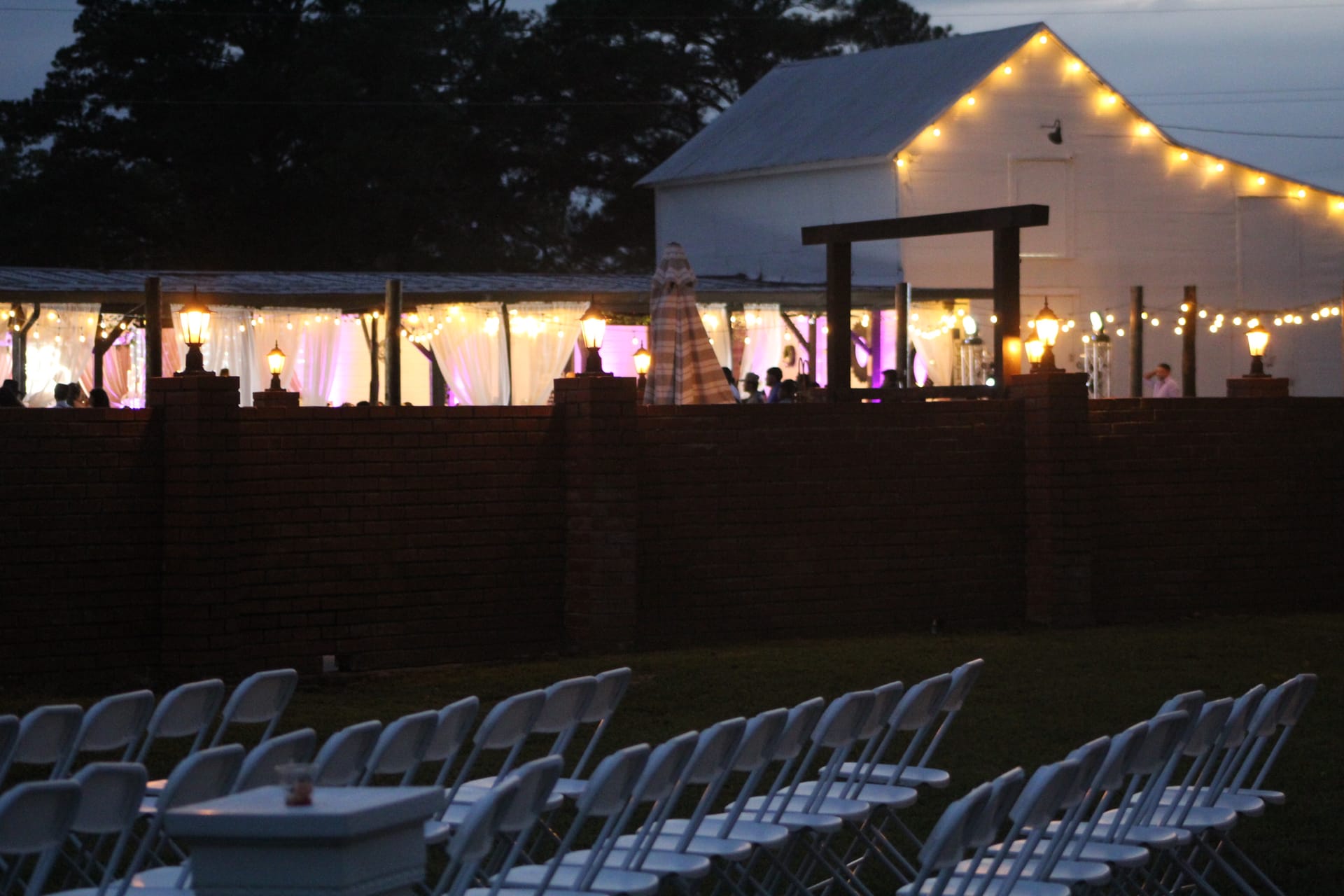 Twilight view of Scarborough House wedding venue with glowing string lights draping the magnolia pavilion and white barn, white folding chairs arranged on the ceremony lawn in the foreground.
