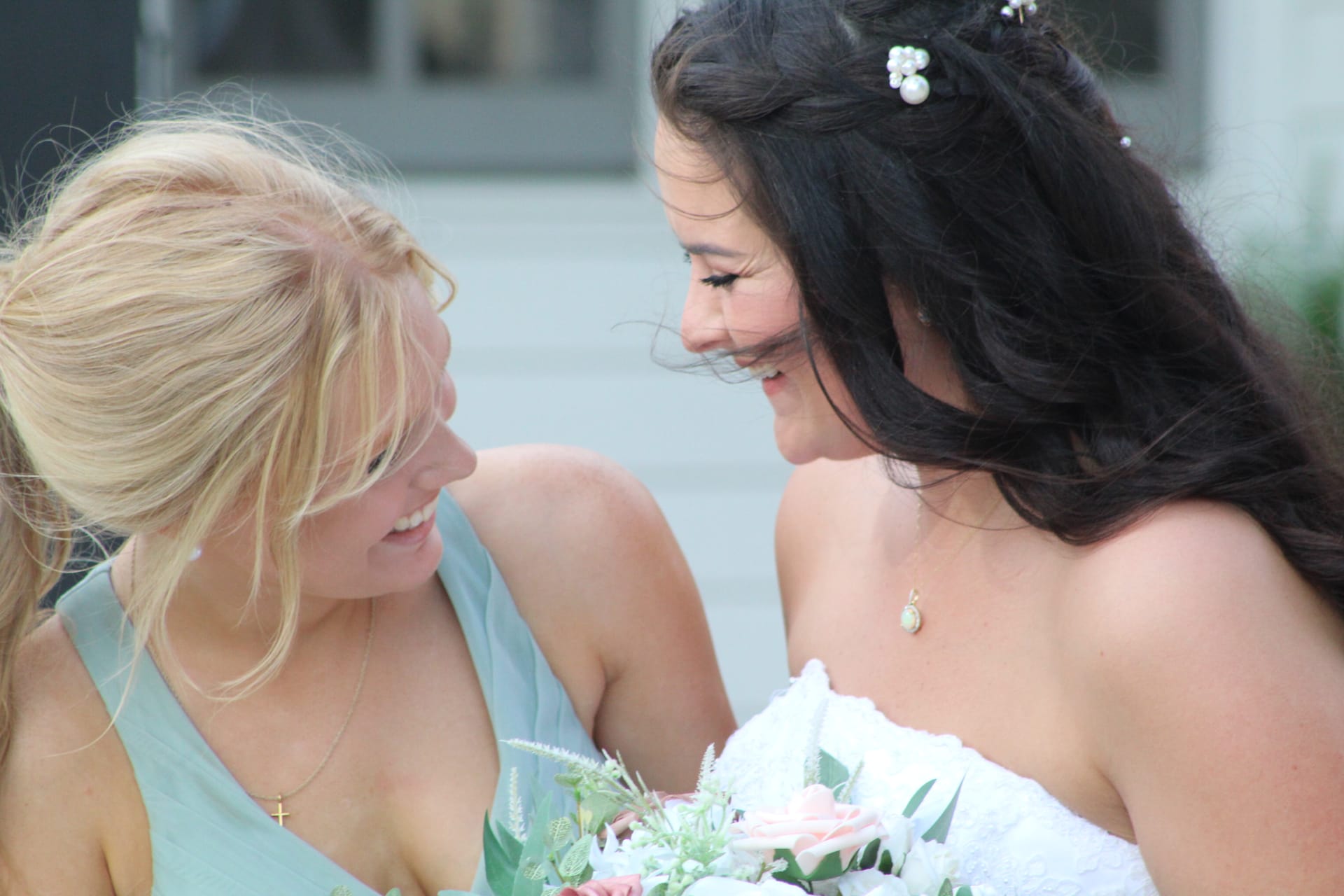 Bride in white strapless gown and bridesmaid in sage green dress share a joyful smile while holding a blush and white floral bouquet at Scarborough House wedding venue.