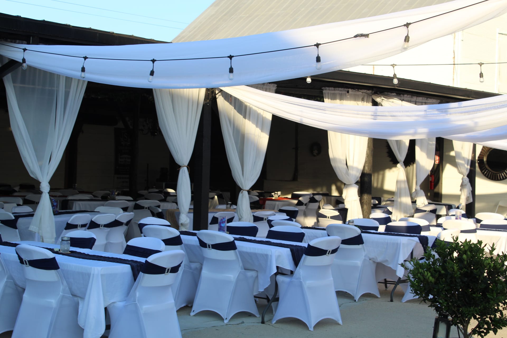 Elegant outdoor reception area draped in white fabric with string lights overhead and round tables set with white linens and navy chair sashes at Scarborough House wedding venue.