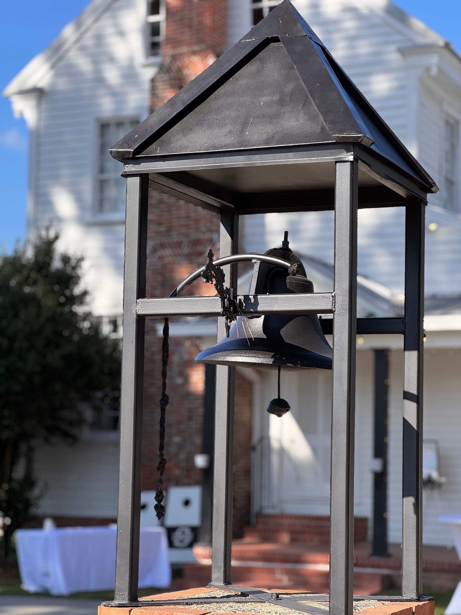 A black iron bell suspended in an ornate metal shelter stands in front of the historic 1821 Scarborough House manor on a sunny day.