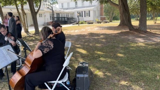 String quartet musicians seated outdoors among oak trees performing at a wedding ceremony with the historic 1821 Scarborough House manor visible in the background