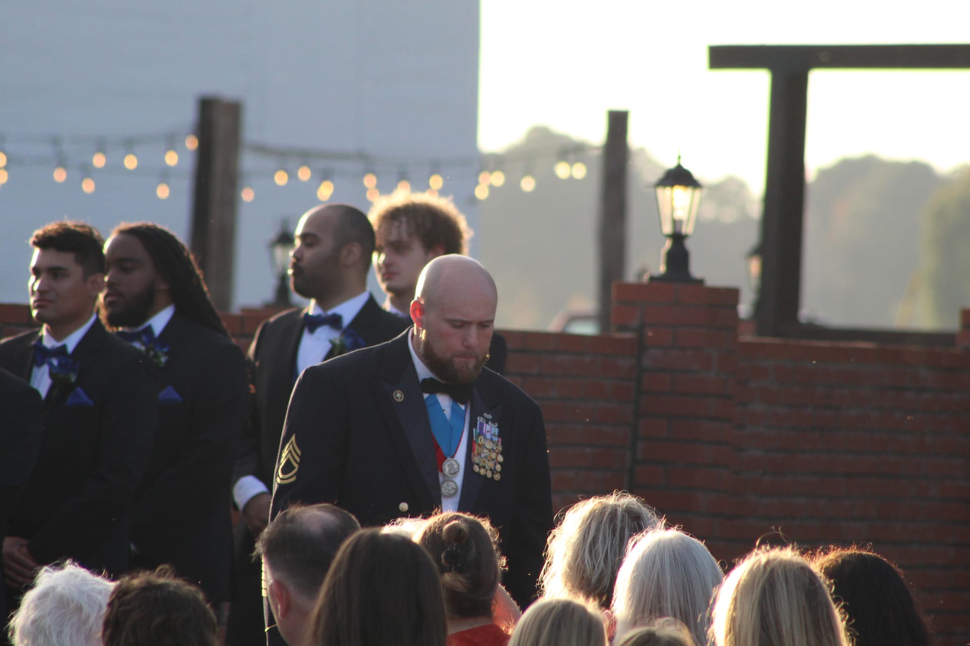 A groomsman in formal attire standing thoughtfully outdoors during an evening wedding ceremony with string lights in the background.