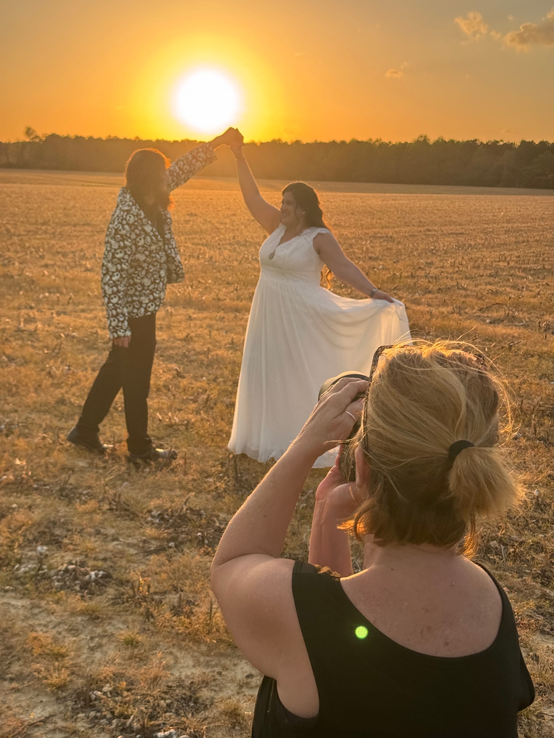 A photographer captures a bride in a flowing white gown twirling with a companion in a golden farmland field at sunset at Scarborough House estate.