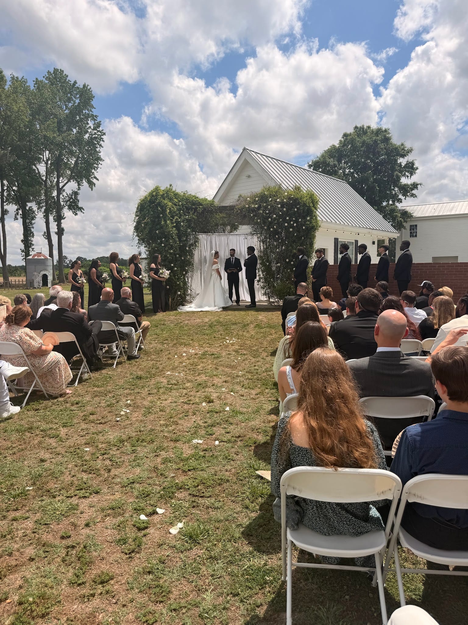 Outdoor wedding ceremony on the lawn at Scarborough House with bride and groom at a floral arch in front of a white historic chapel surrounded by bridesmaids, groomsmen, and seated guests on a sunny day.