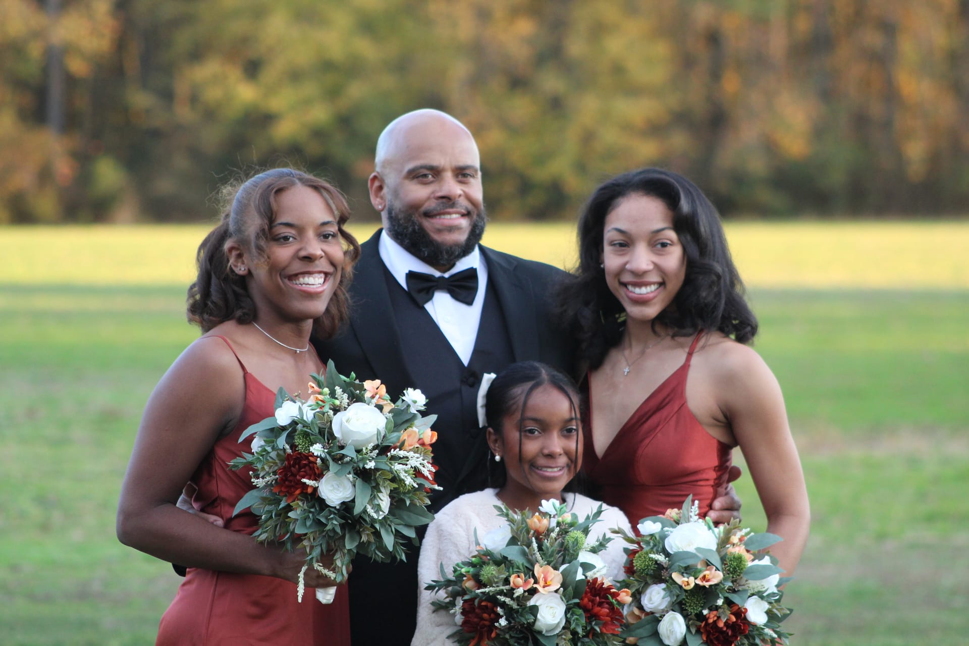 A man in a tuxedo poses with three young women and a girl in burgundy dresses holding autumn floral bouquets on the open farmland fields of Scarborough House estate.