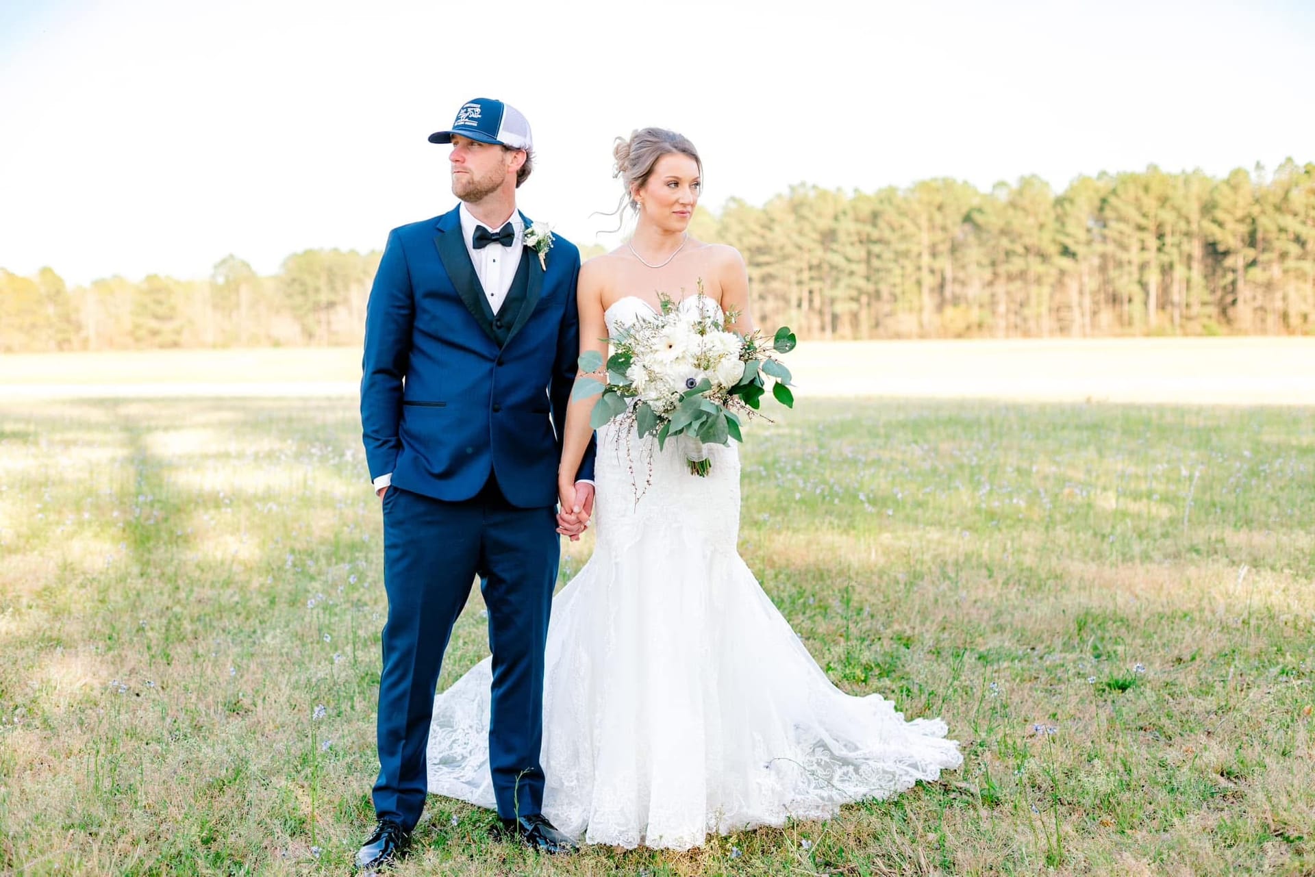Bride in a lace mermaid gown holding a white and greenery bouquet stands hand-in-hand with groom in a navy suit and trucker cap in a sunlit open field at Scarborough House.