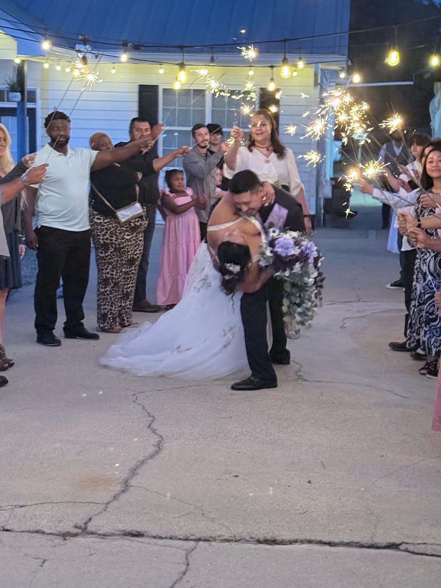 Groom dips and kisses his bride in a romantic moment as wedding guests line the path holding glowing sparklers beneath string lights at an evening outdoor reception.