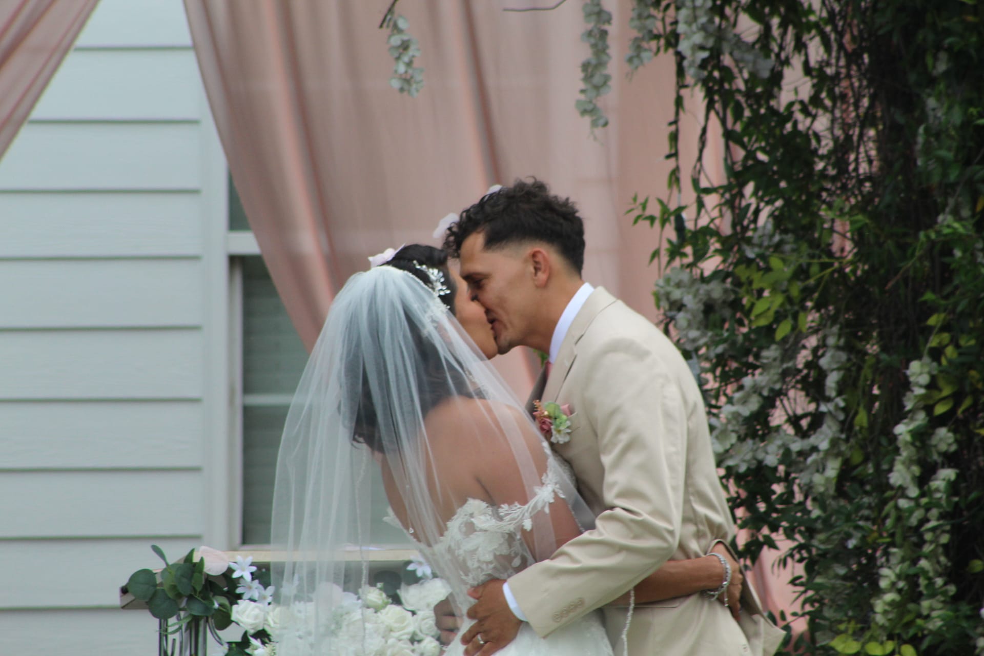 Bride and groom share their first kiss at an outdoor wedding ceremony at Scarborough House, framed by lush greenery and soft pink drapery.