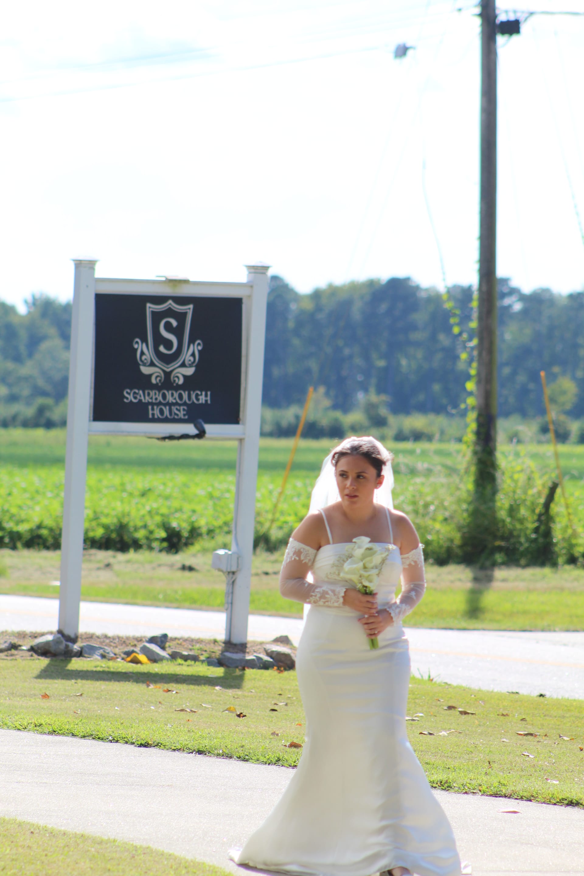 Bride in a fitted white off-shoulder gown holding a white calla lily bouquet stands near the Scarborough House marquee sign with lush green farmland fields in the background.