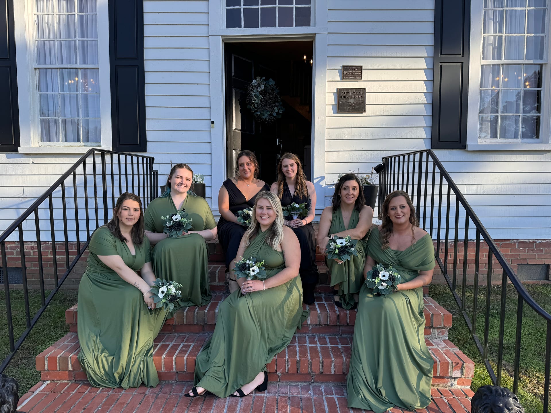 Seven bridesmaids in sage green gowns holding white and navy floral bouquets pose on the brick front steps of the historic 1821 Scarborough House manor.