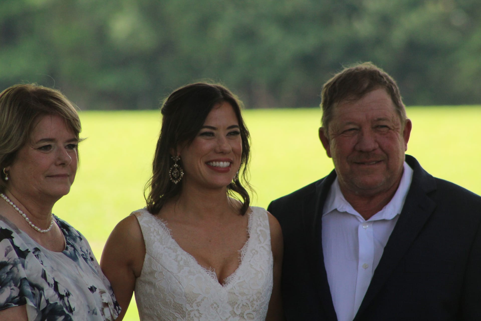 Bride in lace wedding gown smiling with her parents in front of lush green farmland fields at Scarborough House estate.