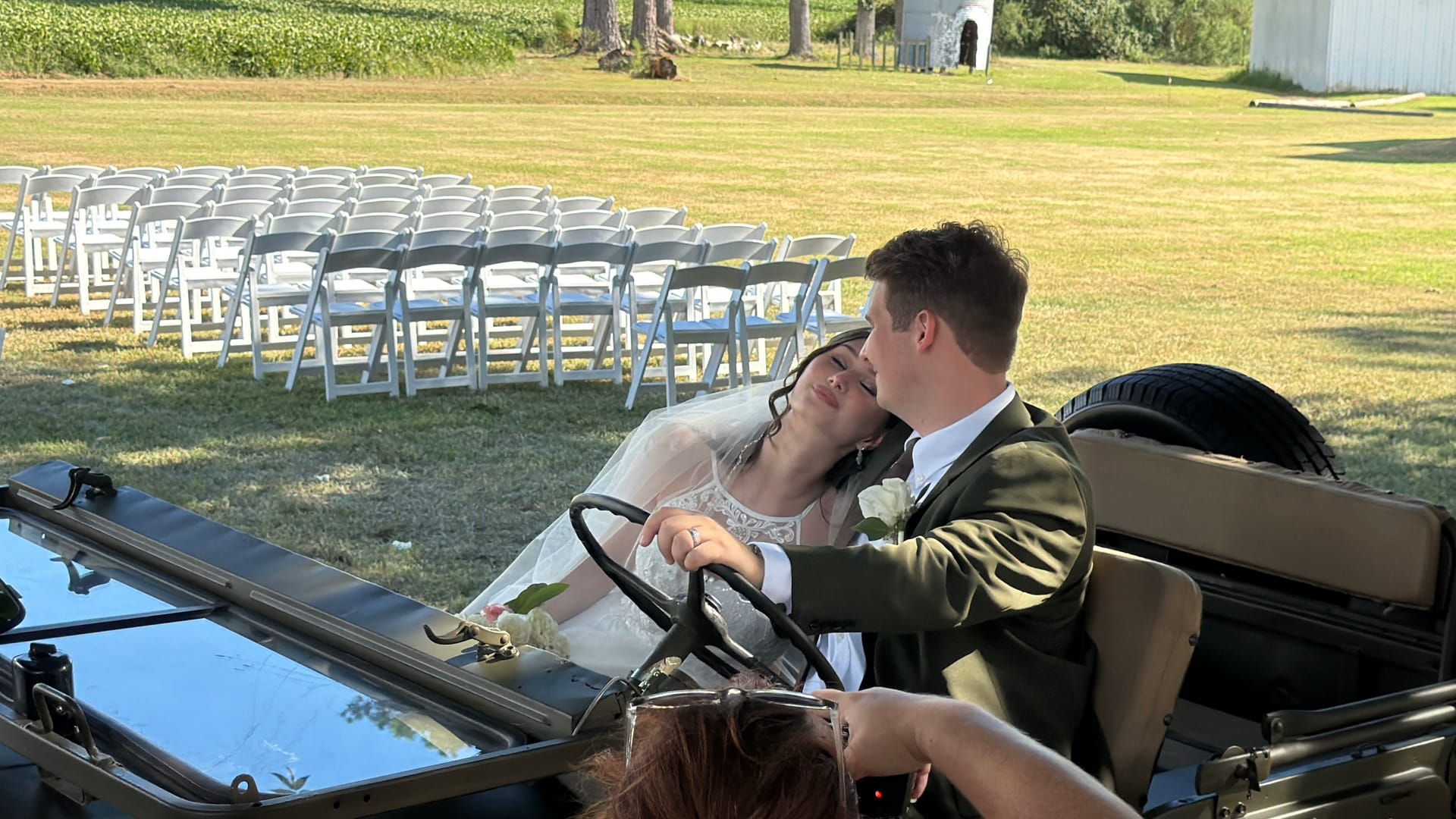 Bride leaning lovingly against the groom as he drives a vintage military jeep across the sunlit ceremony lawn at Scarborough House, with rows of white folding chairs and lush green fields in the background.
