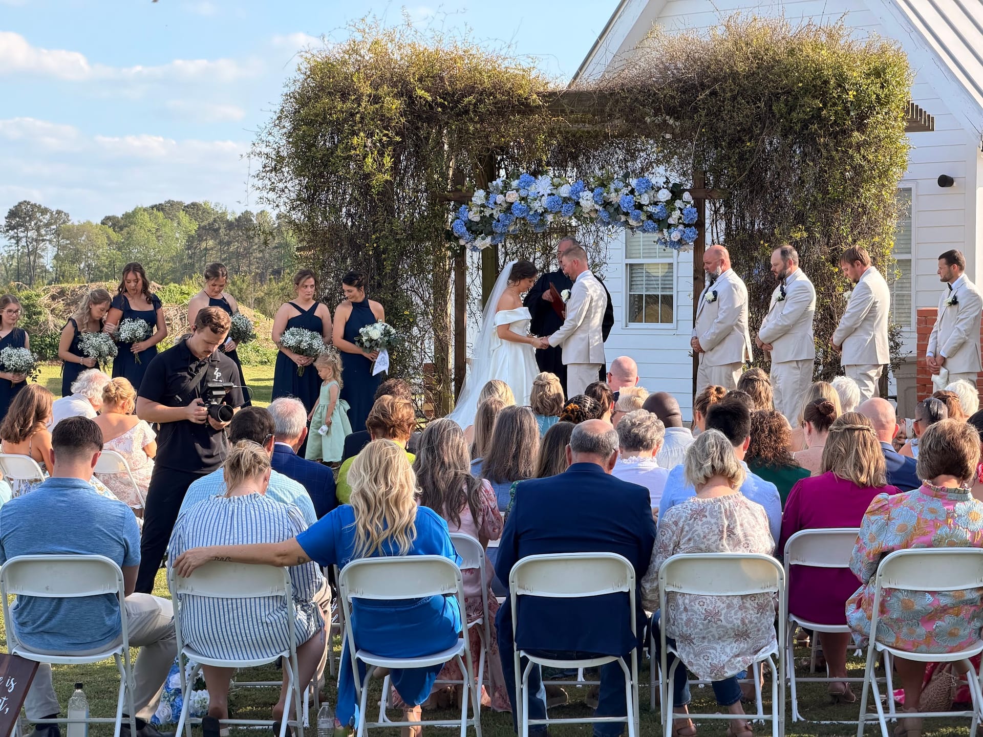 Outdoor wedding ceremony on the lawn at Scarborough House with bride and groom exchanging vows beneath a lush greenery and blue hydrangea arch surrounded by bridesmaids in navy and groomsmen in tan suits before a white historic chapel