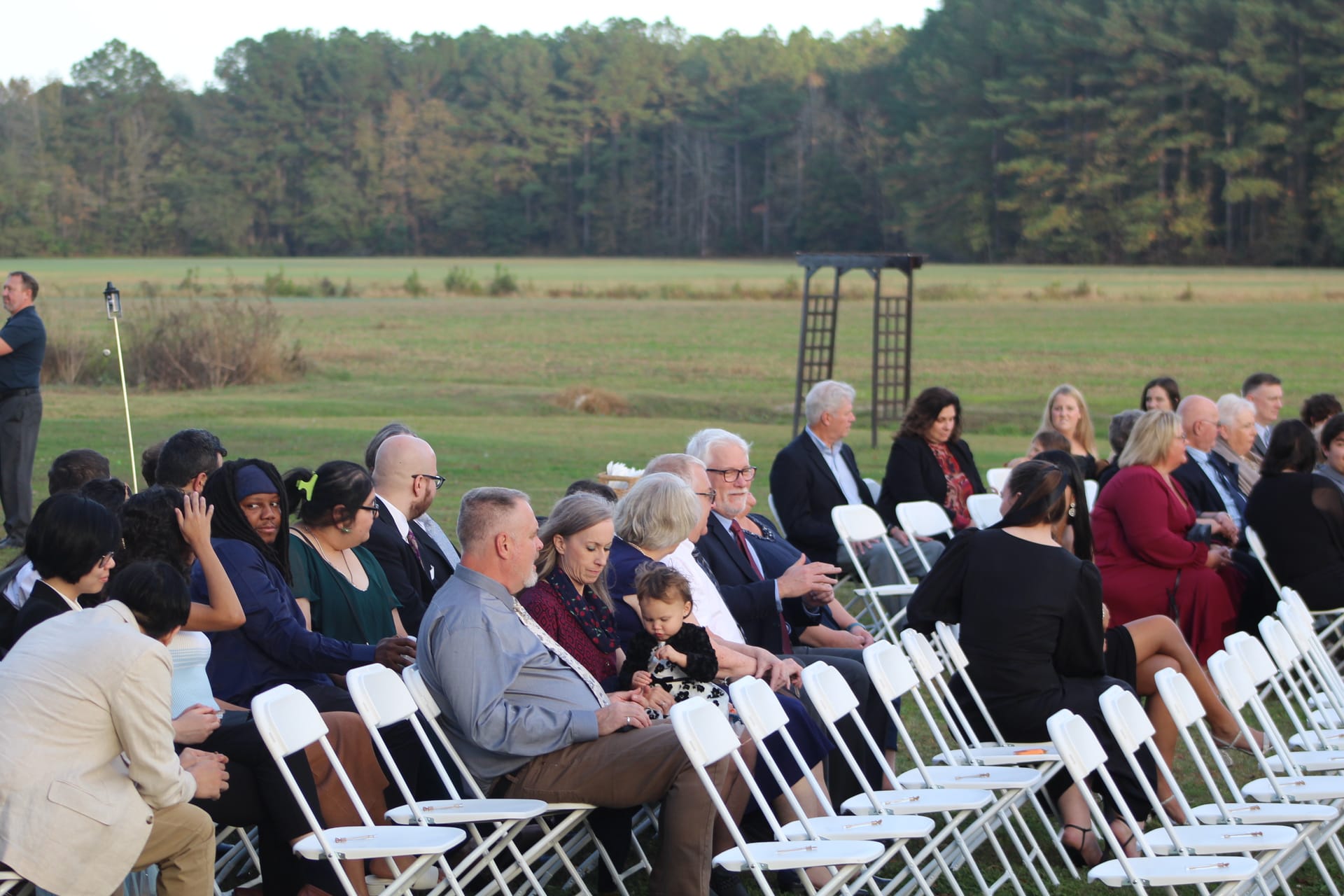 Wedding guests seated in white folding chairs on the ceremony lawn at Scarborough House with open farmland fields and tree line in the background.