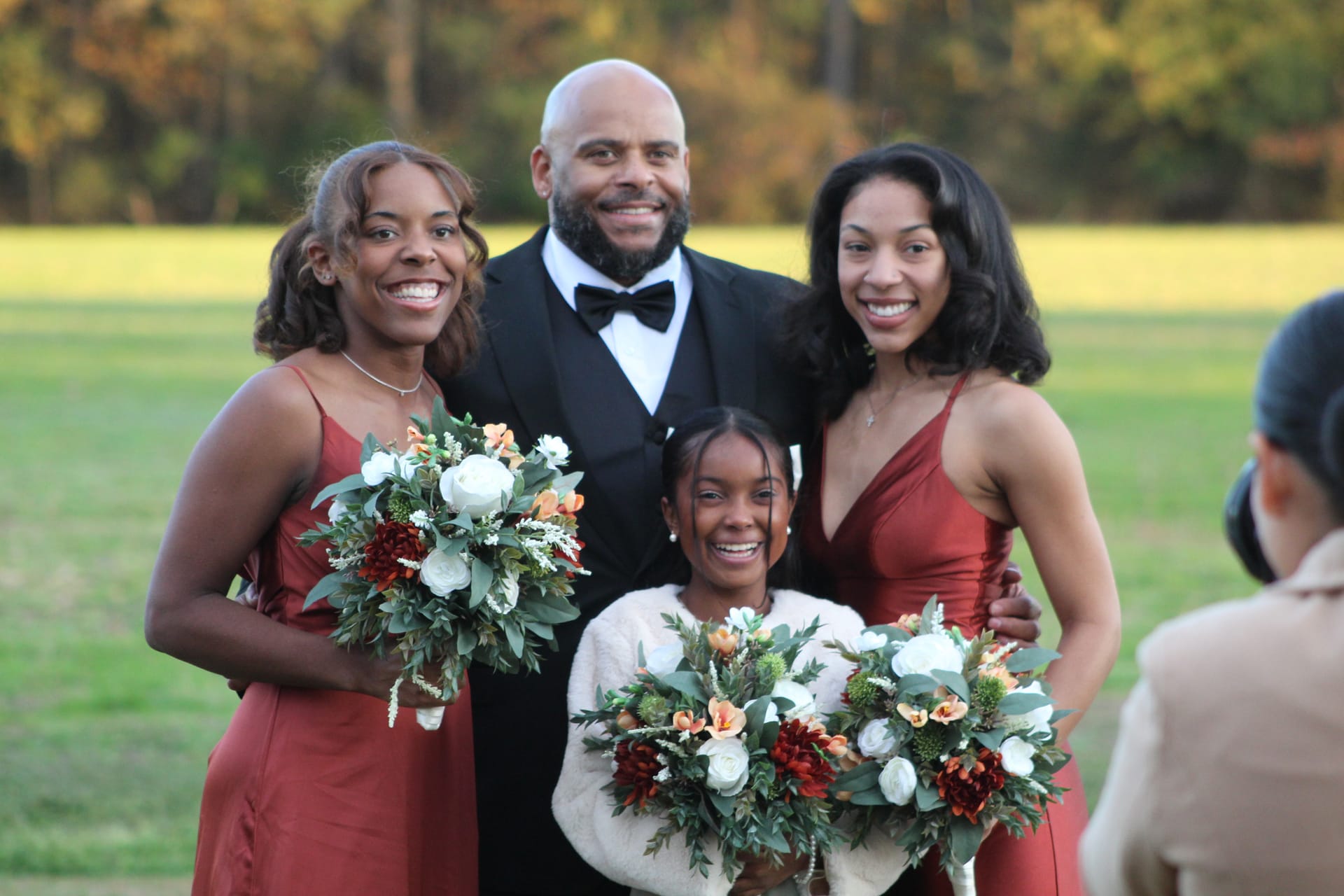 A man in a black tuxedo poses with three bridesmaids in rust-colored dresses holding autumn floral bouquets on the open lawn at Scarborough House with fall foliage in the background.