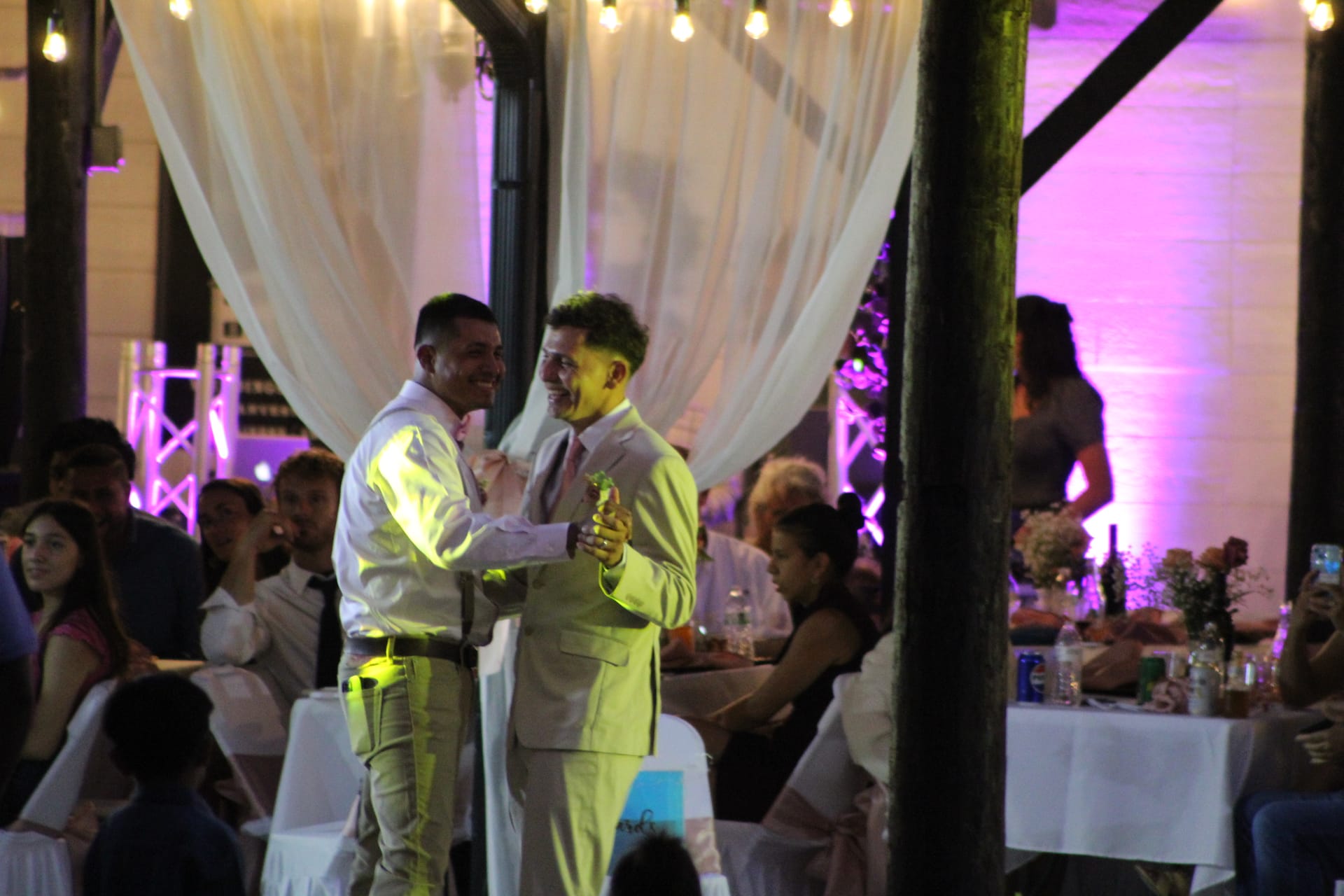 Two grooms smile joyfully as they share their first dance in a warmly lit reception hall decorated with draped white fabric and string lights while guests look on.