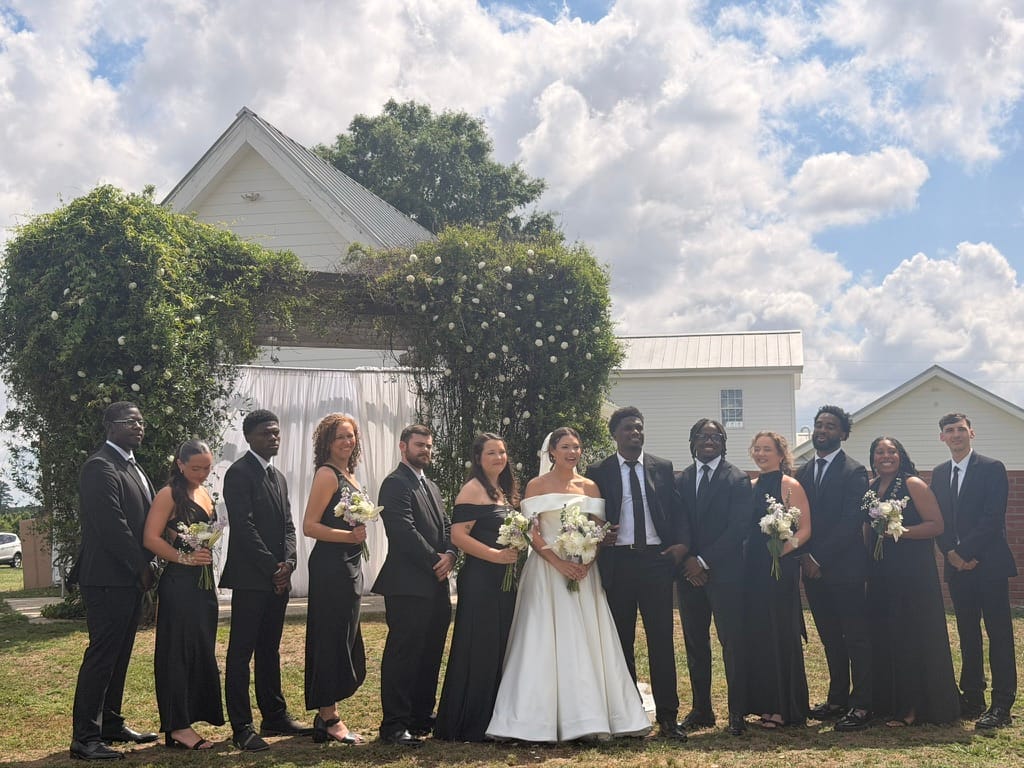 Bride in an off-shoulder white gown stands with her full wedding party dressed in black against a floral greenery arch and white historic chapel at Scarborough House.