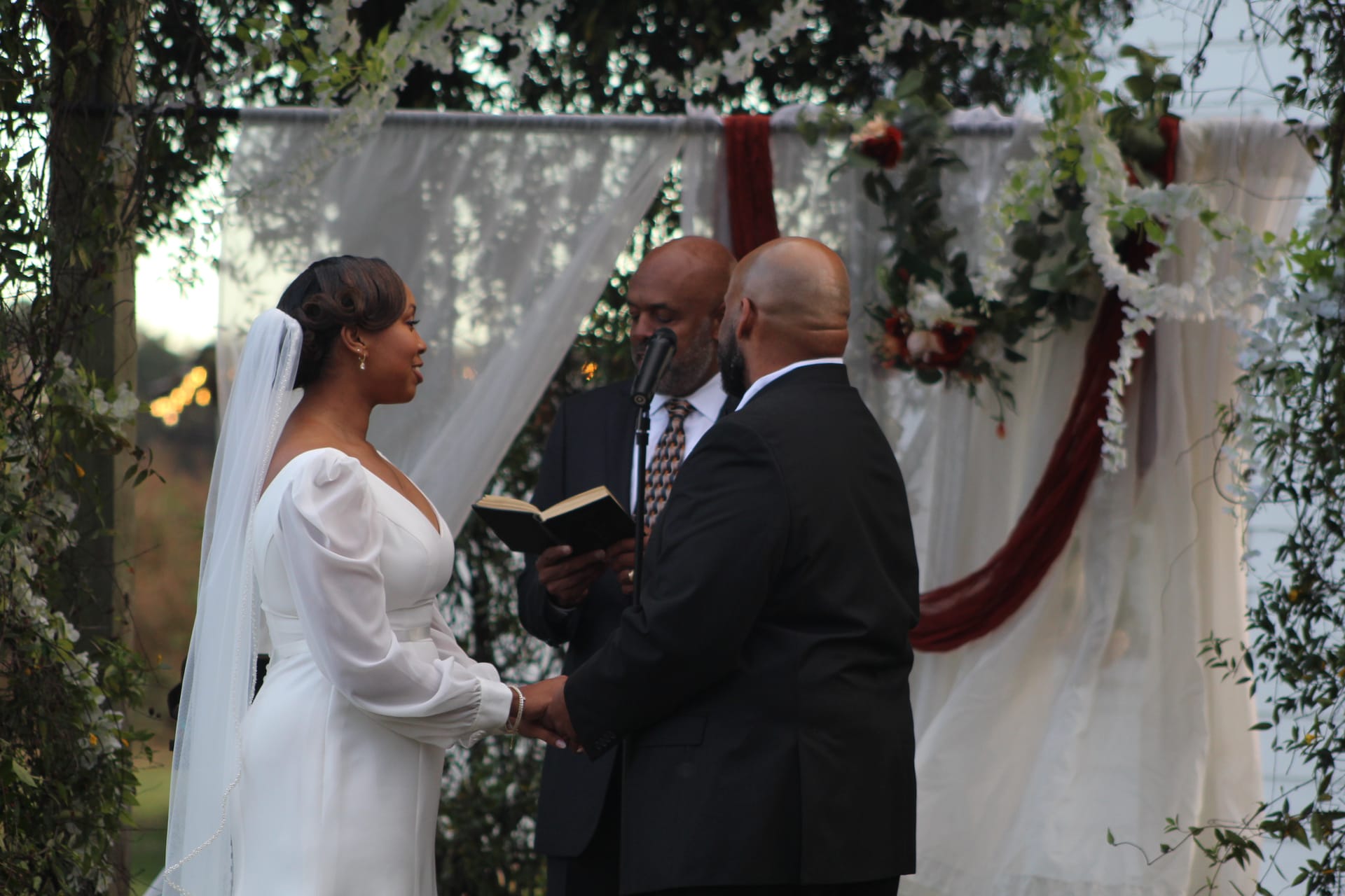 Bride in a white long-sleeve gown and veil holds hands with her groom during outdoor wedding vows beneath a floral and draped arch adorned with white flowers and burgundy fabric at Scarborough House.