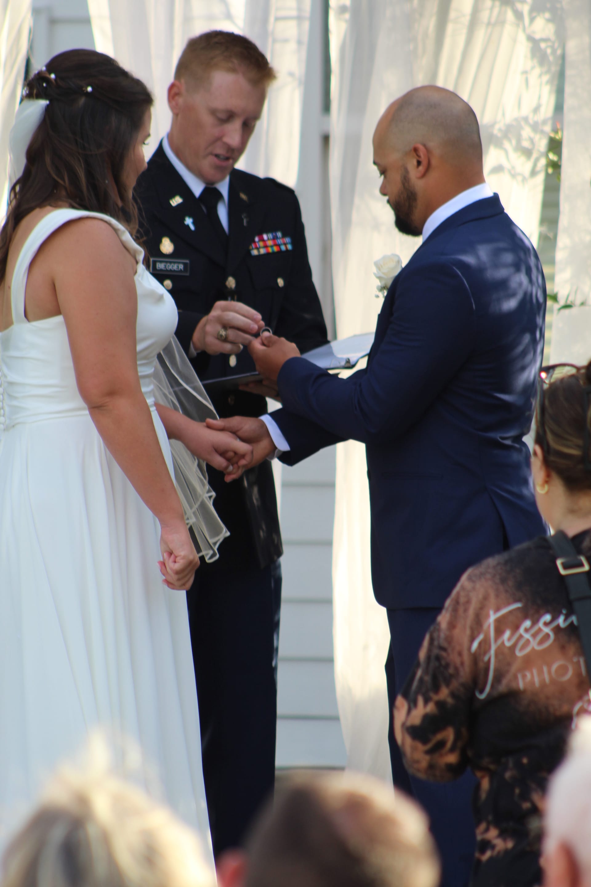 Groom places wedding ring on bride's finger during outdoor ceremony officiated by a military officer at Scarborough House historic estate.