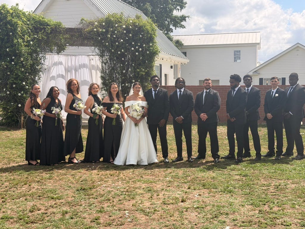 Bride in white off-shoulder gown flanked by bridesmaids in black dresses and groomsmen in black suits posed on the ceremony lawn in front of a floral arch at Scarborough House.
