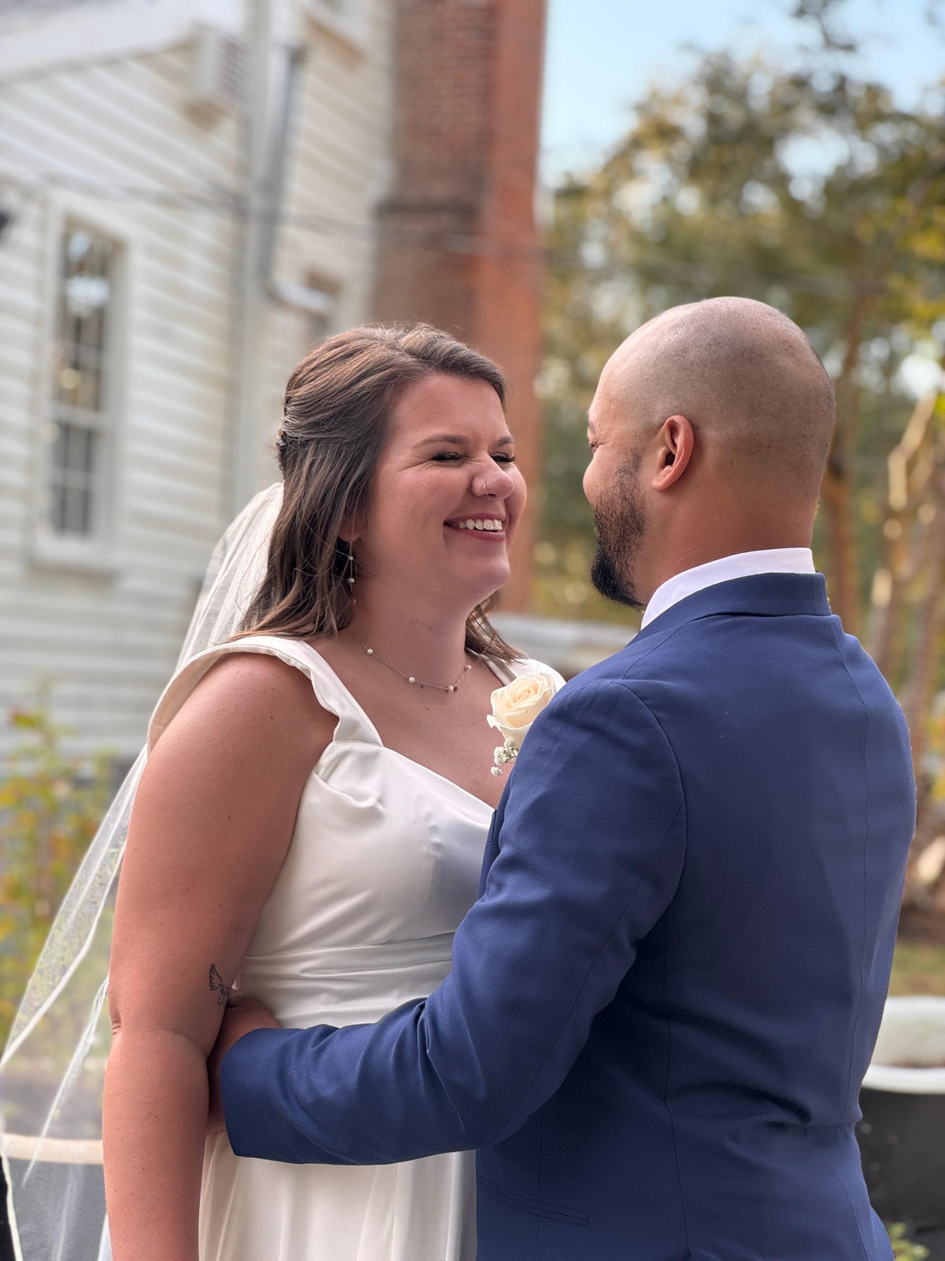 A smiling bride in a white gown and veil gazes joyfully at her groom in a navy suit during their outdoor wedding ceremony in front of the historic 1821 Scarborough House manor.
