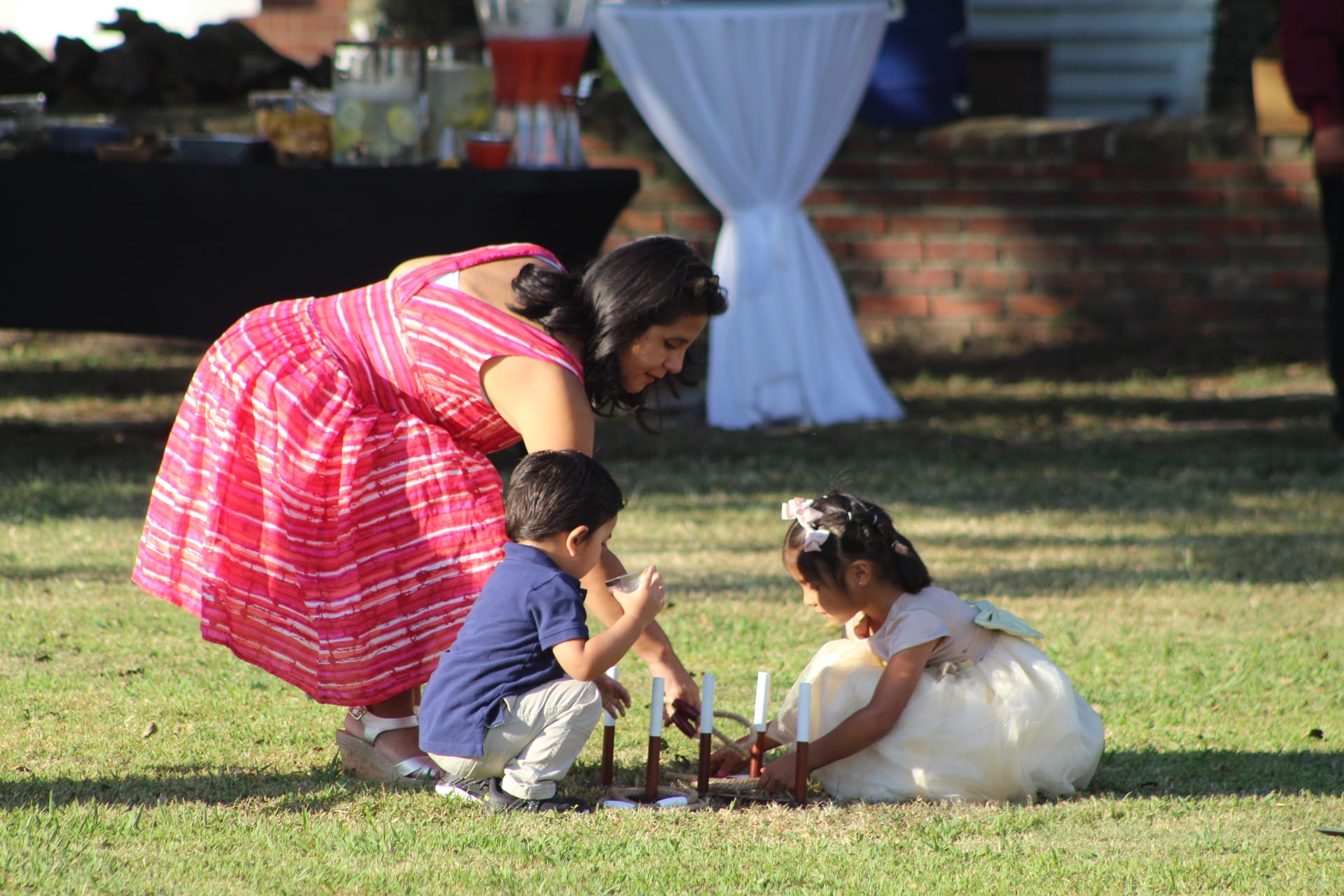 A woman in a pink dress bends down to help two young children arrange candles on the grass at an outdoor wedding reception at Scarborough House.