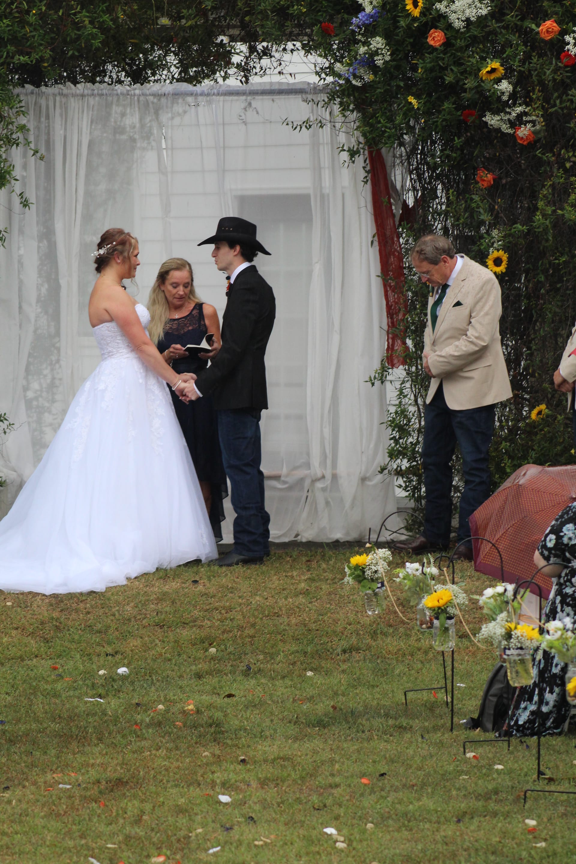 Bride in white ball gown and groom in black cowboy hat exchange vows beneath a flower-adorned arch on the ceremony lawn at Scarborough House historic estate.