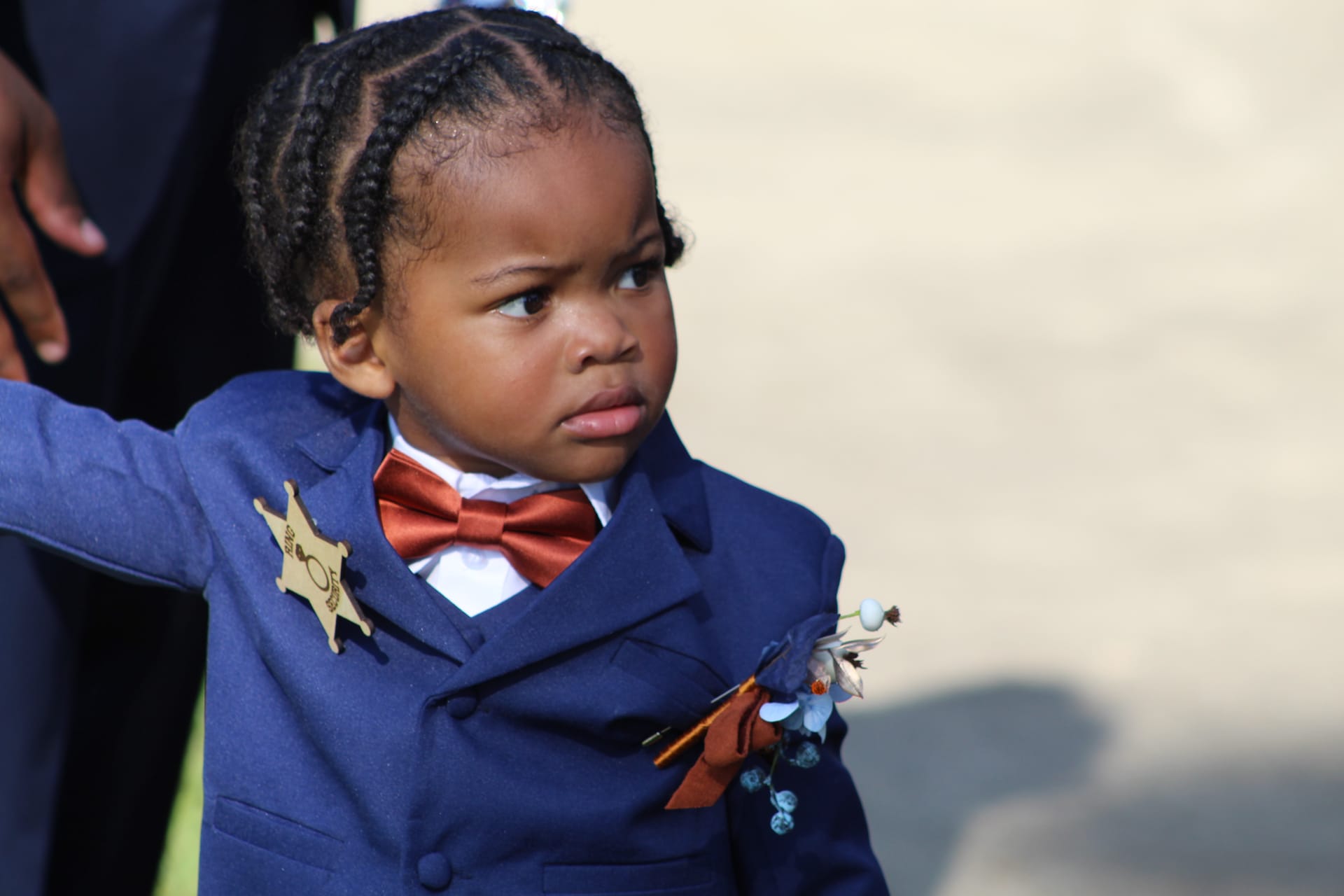 A young ring bearer dressed in a navy suit with a rust bow tie and floral boutonniere gazes to the side during an outdoor wedding ceremony at Scarborough House.