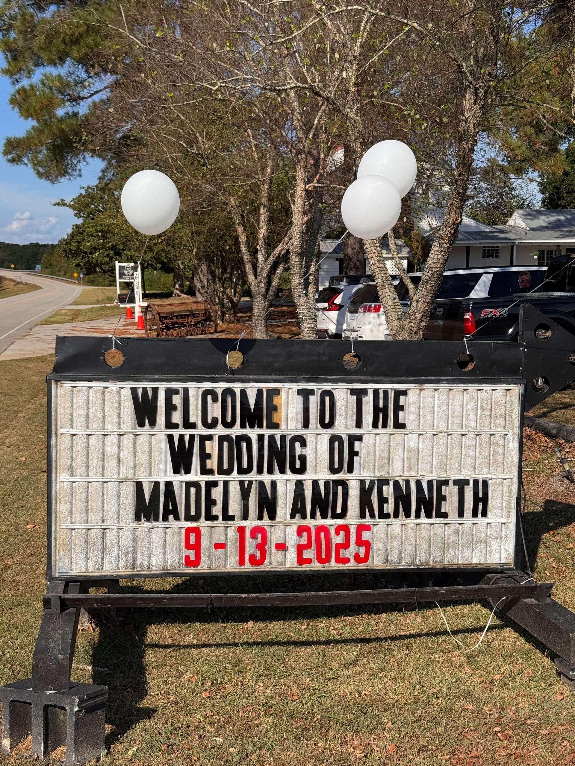 Outdoor marquee sign decorated with white balloons welcoming guests to the wedding of Madelyn and Kenneth on September 13 2025 at a tree-lined estate venue.