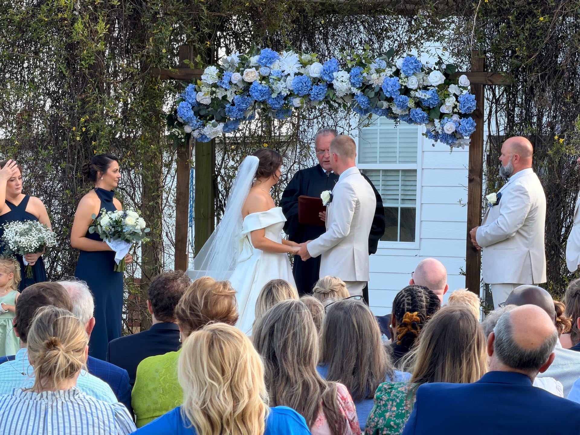 Bride and groom exchange vows beneath a lush blue and white hydrangea floral arch at an outdoor wedding ceremony surrounded by guests and the wedding party.