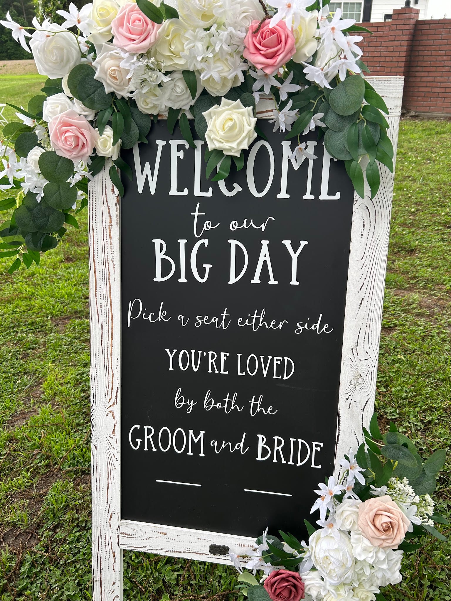 A rustic chalkboard wedding welcome sign reading Welcome to our Big Day framed with pink and white roses and greenery on the ceremony lawn at Scarborough House.