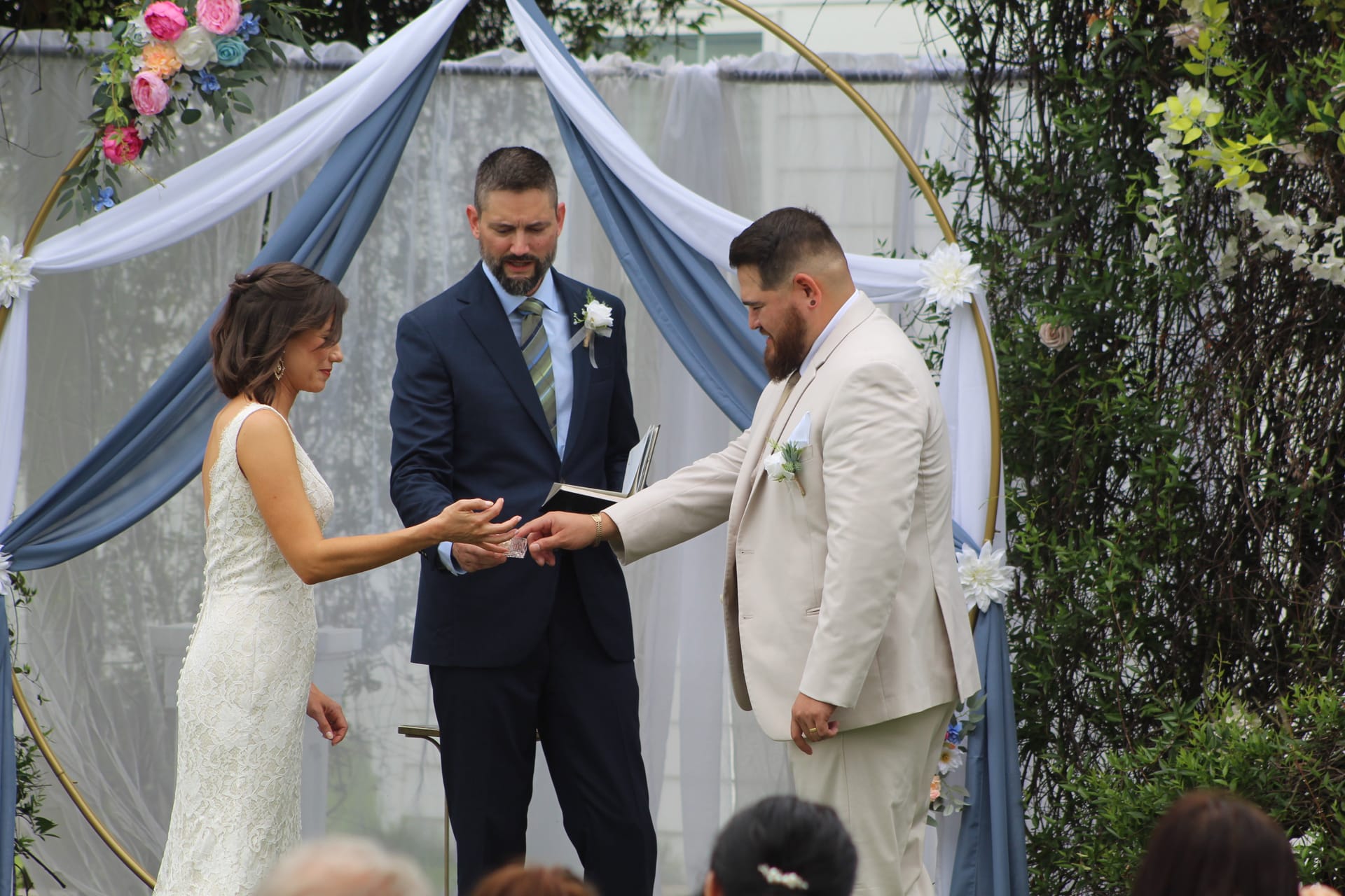 Bride placing a wedding ring on the grooms finger during an outdoor ceremony beneath a gold circular arch adorned with blue drapery and colorful florals.