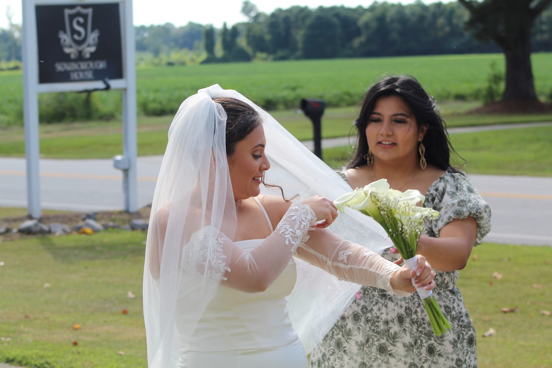 Bride in white gown and veil smiling as bridesmaid hands her a white calla lily bouquet outside the Scarborough House estate sign on a sunny wedding day.