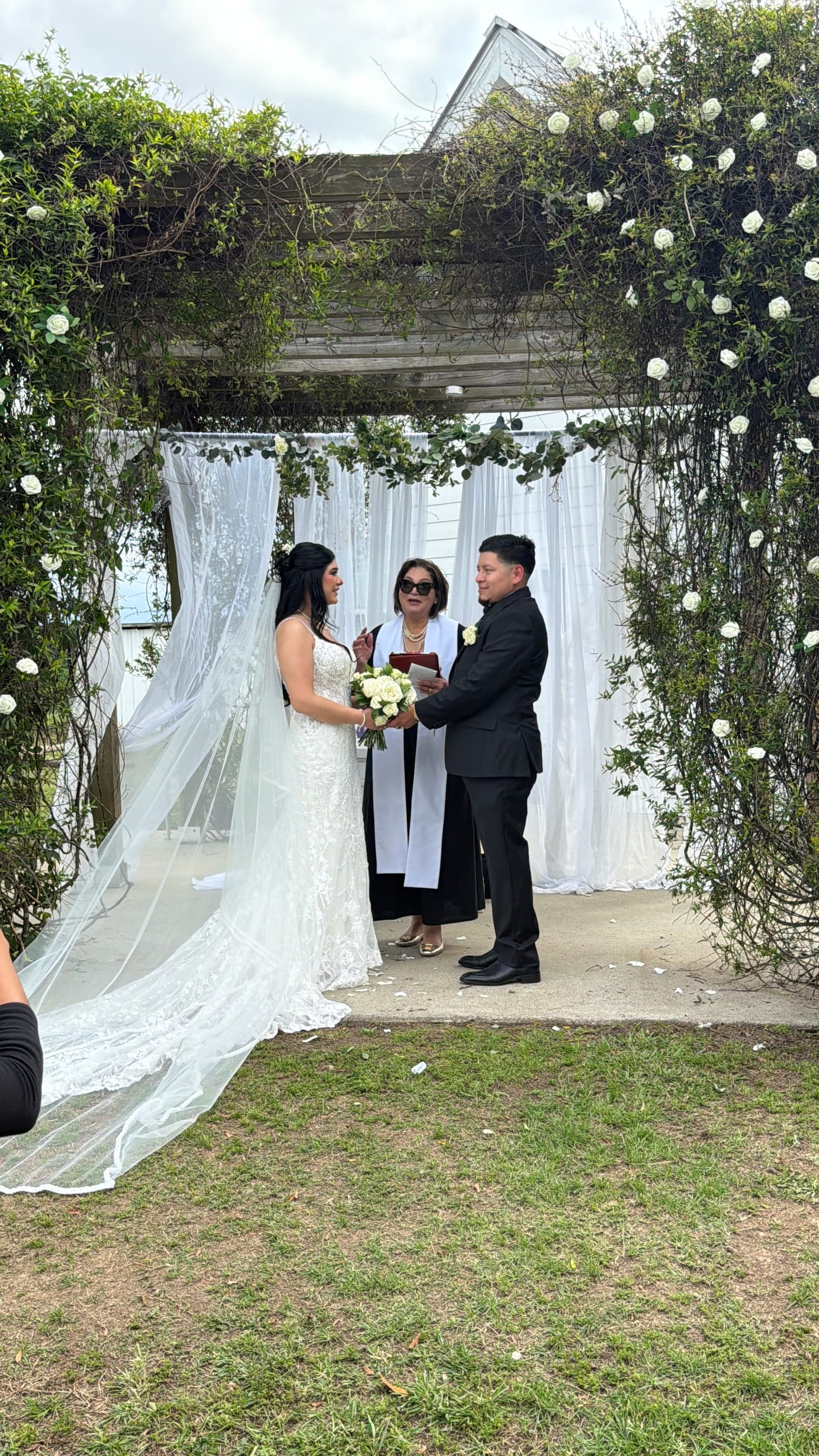Bride in a lace gown and groom in a black suit exchange vows beneath a flowering vine arch adorned with white roses and draped white fabric at Scarborough House.