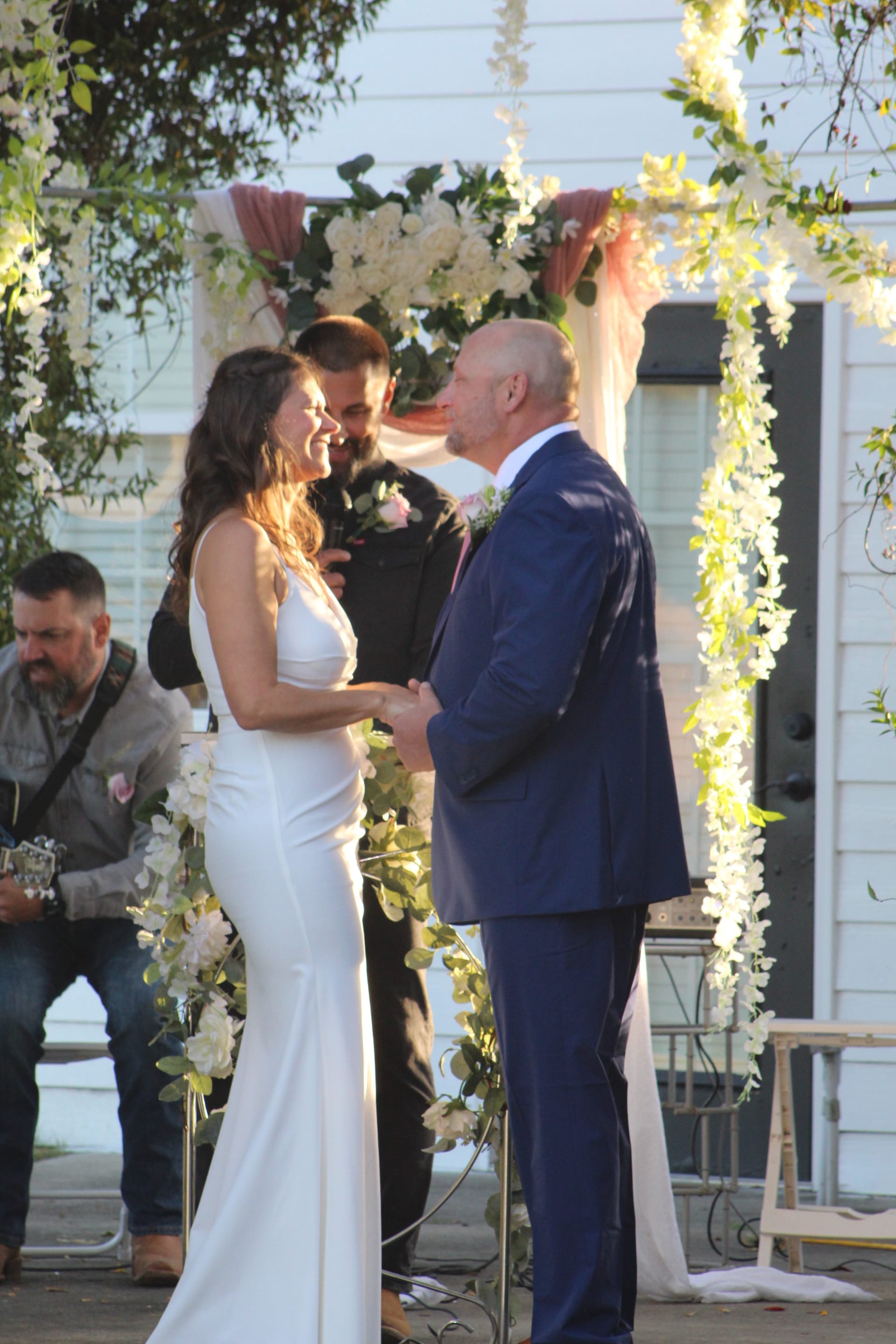Bride in a sleek white gown smiles joyfully at her groom in a navy suit as they exchange vows beneath a flower-draped arch at a golden-hour outdoor wedding ceremony.