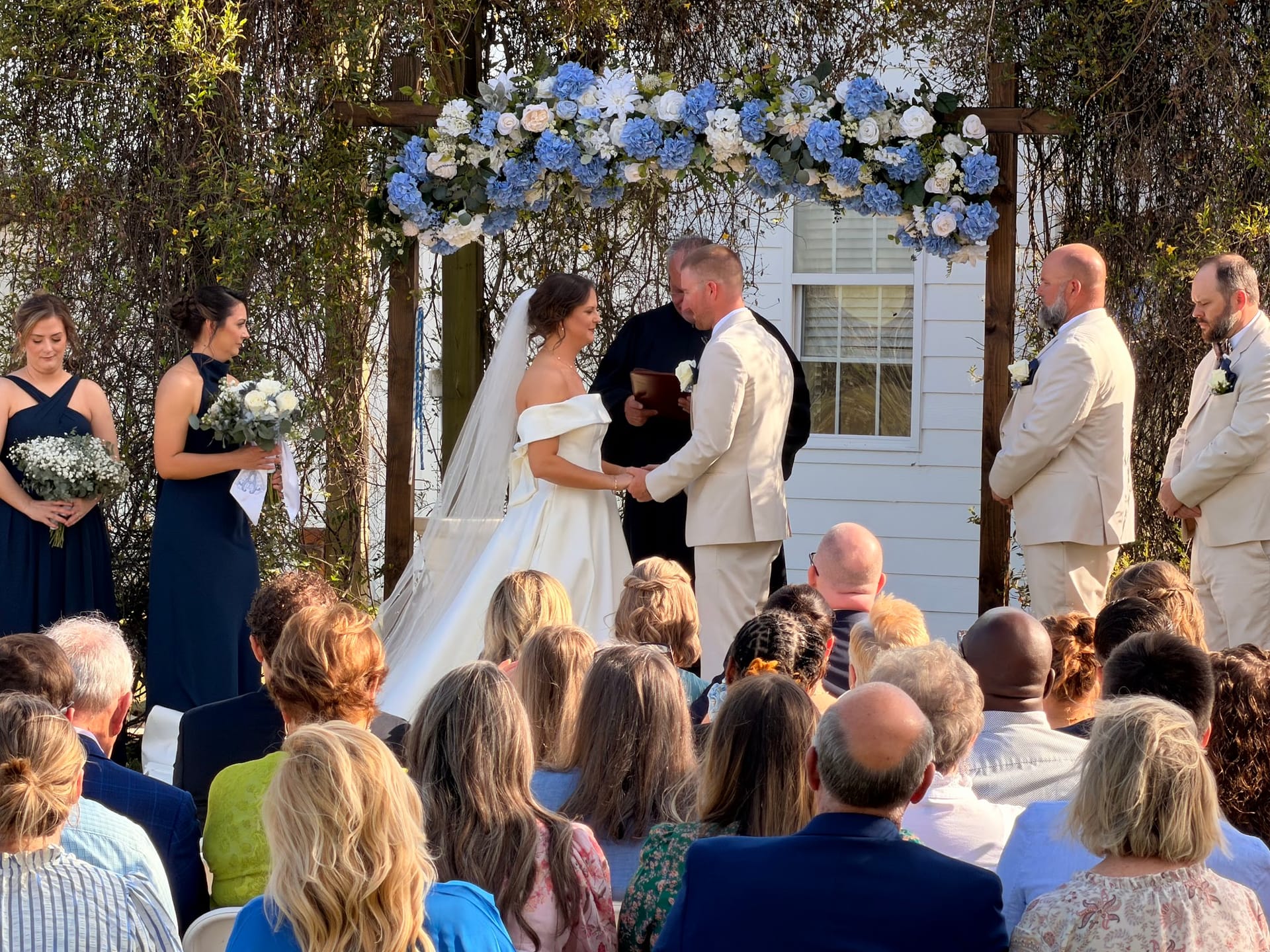 Bride and groom exchange vows beneath a wooden arch adorned with blue hydrangeas and white flowers at an outdoor wedding ceremony while guests and the wedding party look on.