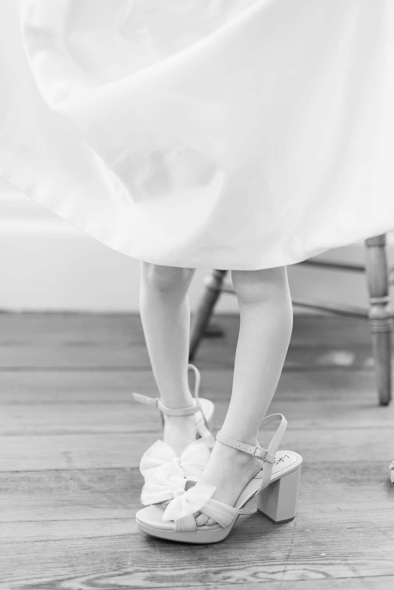 Black and white close-up of a bride wearing white block-heel sandals with bow detail beneath her flowing wedding gown on a wooden floor.