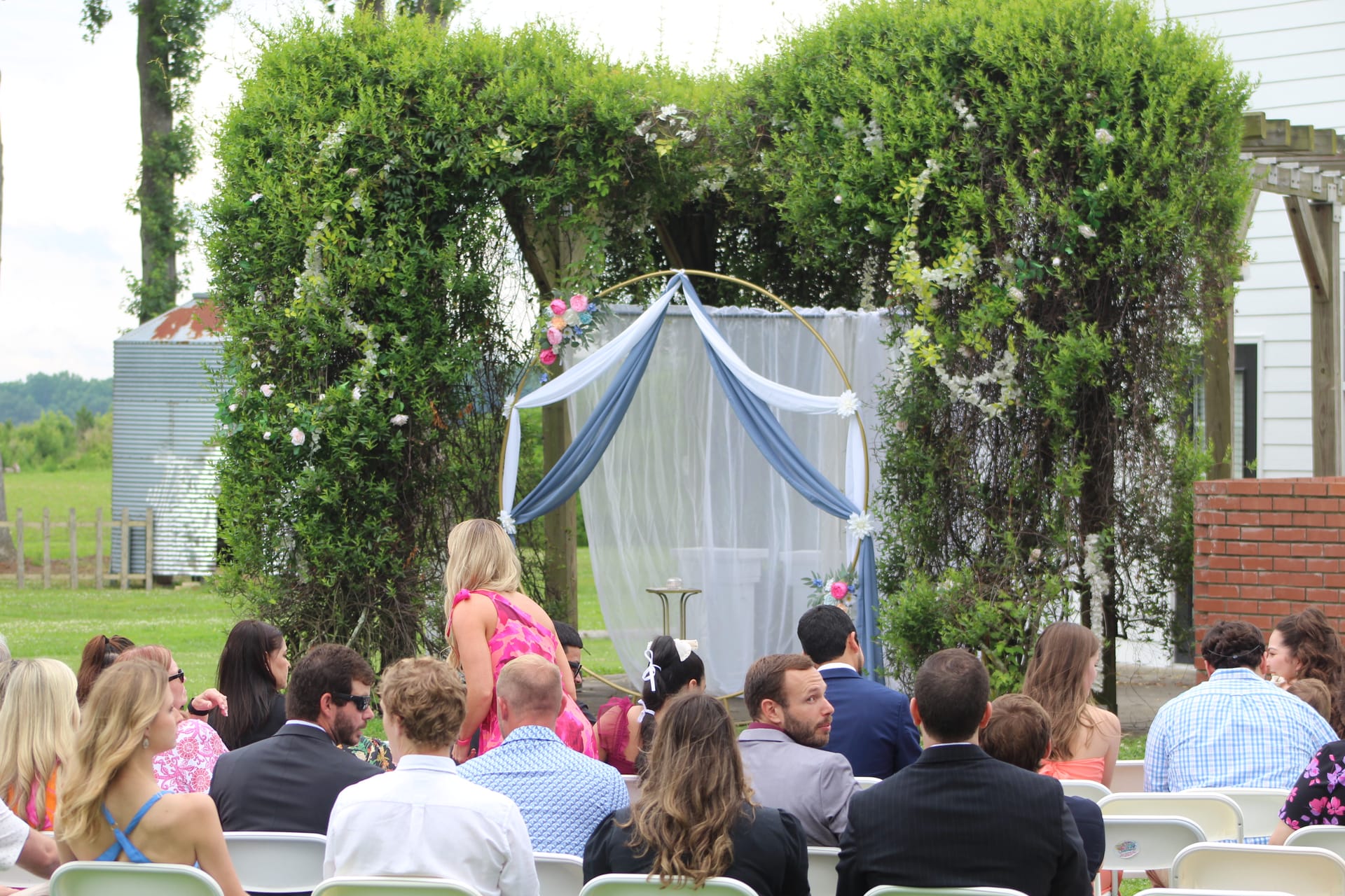 Wedding guests seated in white chairs facing an elegant circular arch draped in blue and white fabric with pink floral accents, framed by sculpted green hedges on the ceremony lawn at Scarborough House.