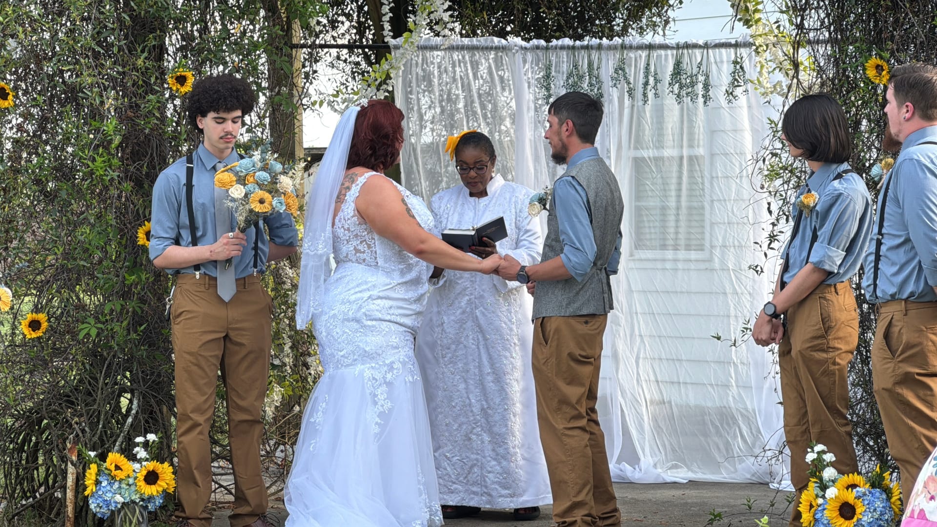 Bride and groom exchange vows at an outdoor ceremony beneath a floral arch decorated with sunflowers, surrounded by groomsmen in blue shirts and tan pants.