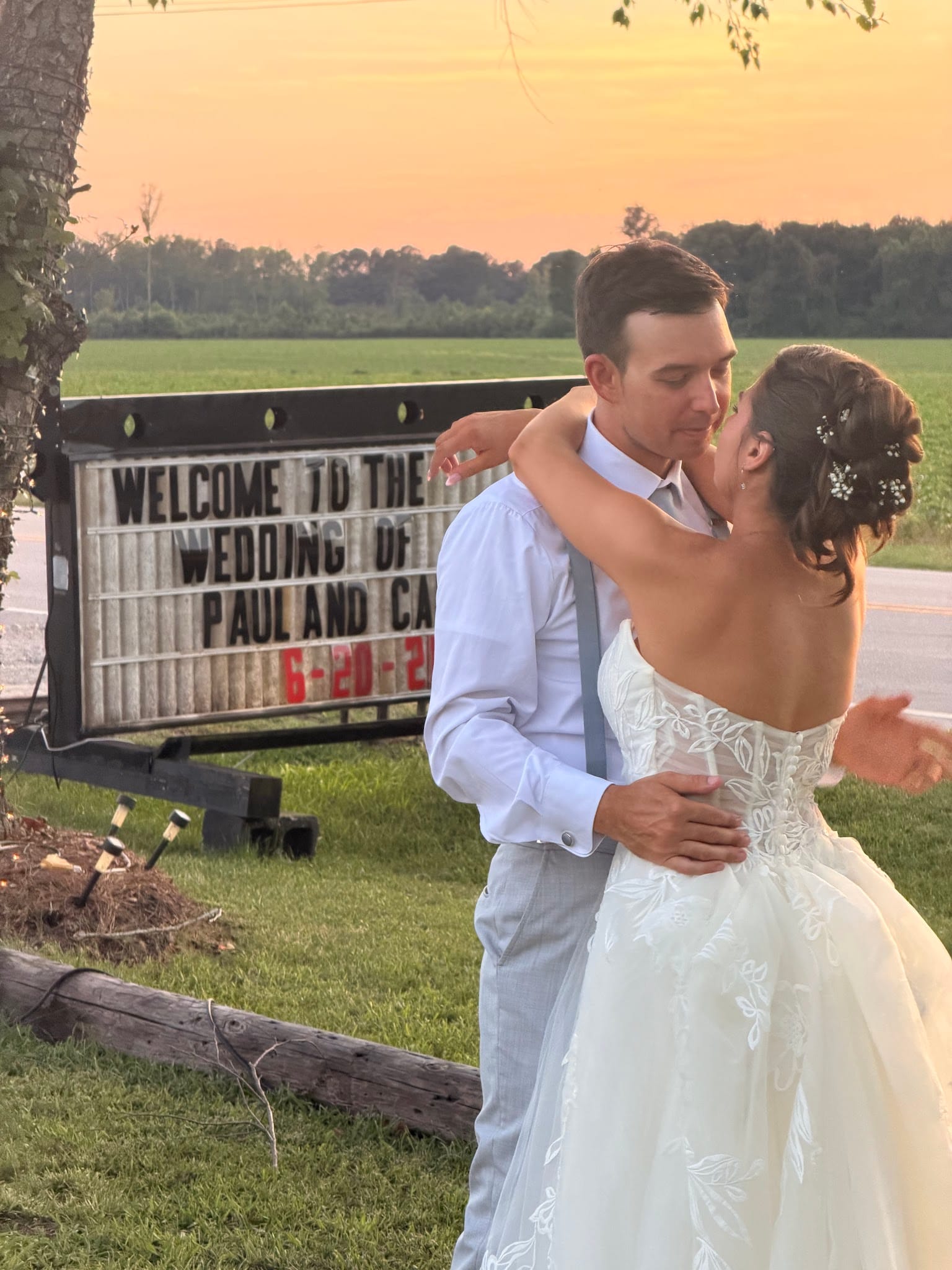 Bride and groom share a tender embrace near the wedding welcome marquee sign at sunset, with open farmland fields glowing in warm golden-hour light behind them.