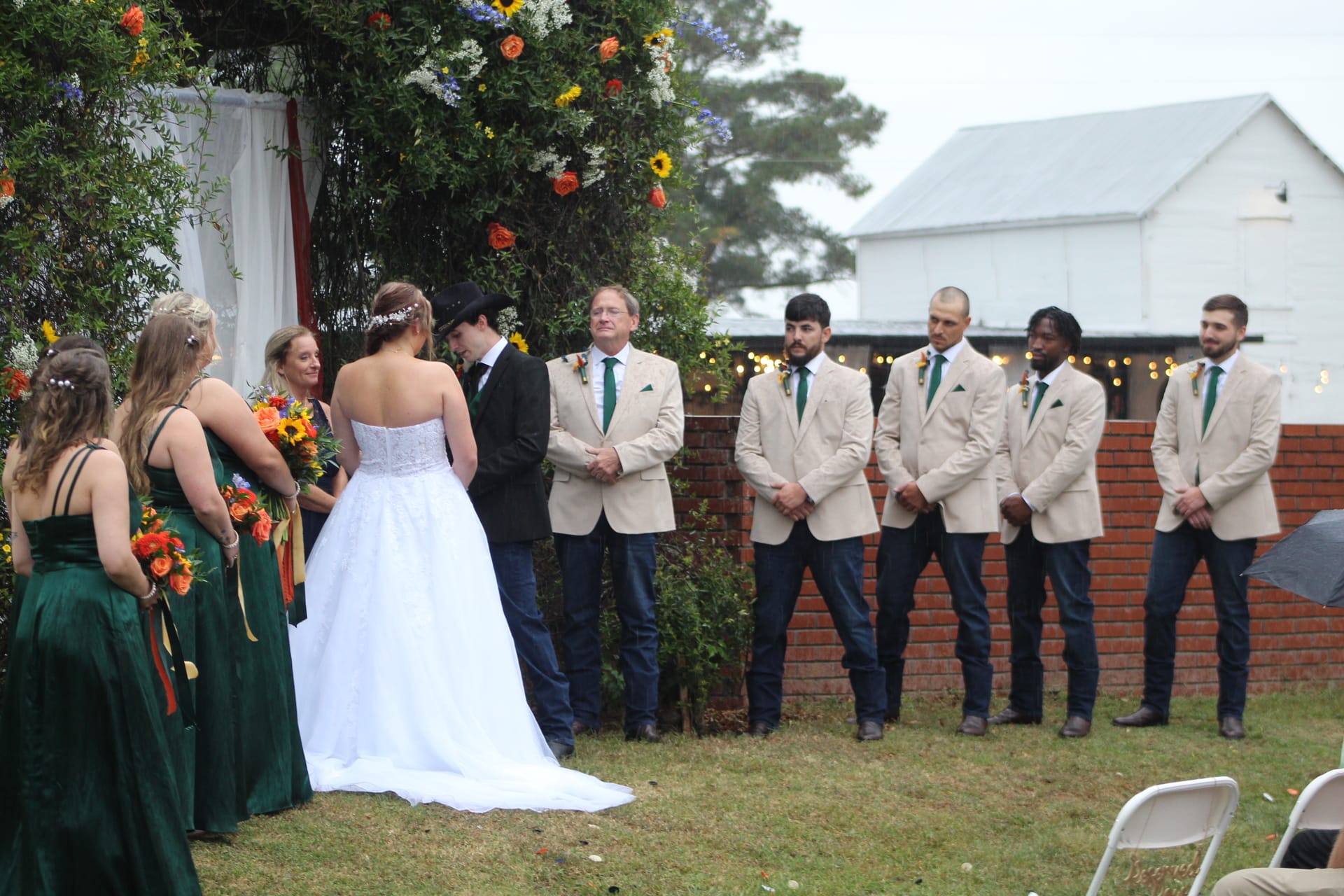 Wedding party stands in formation during an outdoor ceremony beneath a floral arch at Scarborough House, with bridesmaids in emerald green gowns and groomsmen in tan blazers with green ties flanking the bride and groom.