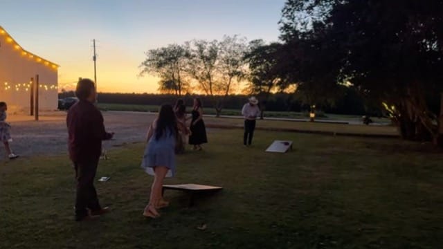 Wedding guests playing cornhole on the lawn at sunset with string lights and a white barn in the background at Scarborough House estate venue.