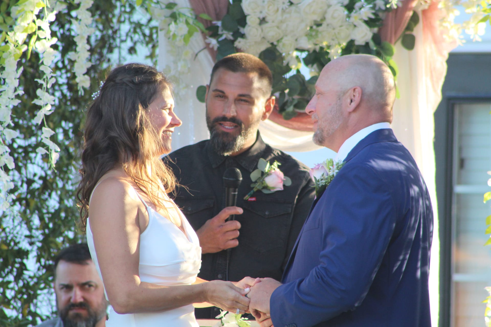 Smiling bride in white gown and groom in navy suit hold hands during their outdoor wedding ceremony beneath a lush floral arch at Scarborough House.
