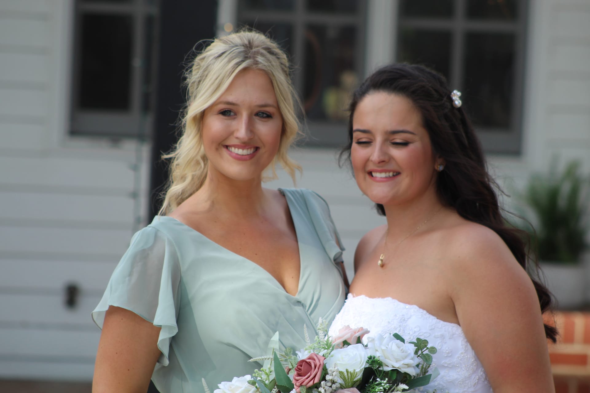A smiling bridesmaid in a sage green dress stands beside a bride in a strapless white gown holding a bouquet of blush and white roses outside a historic white manor house.