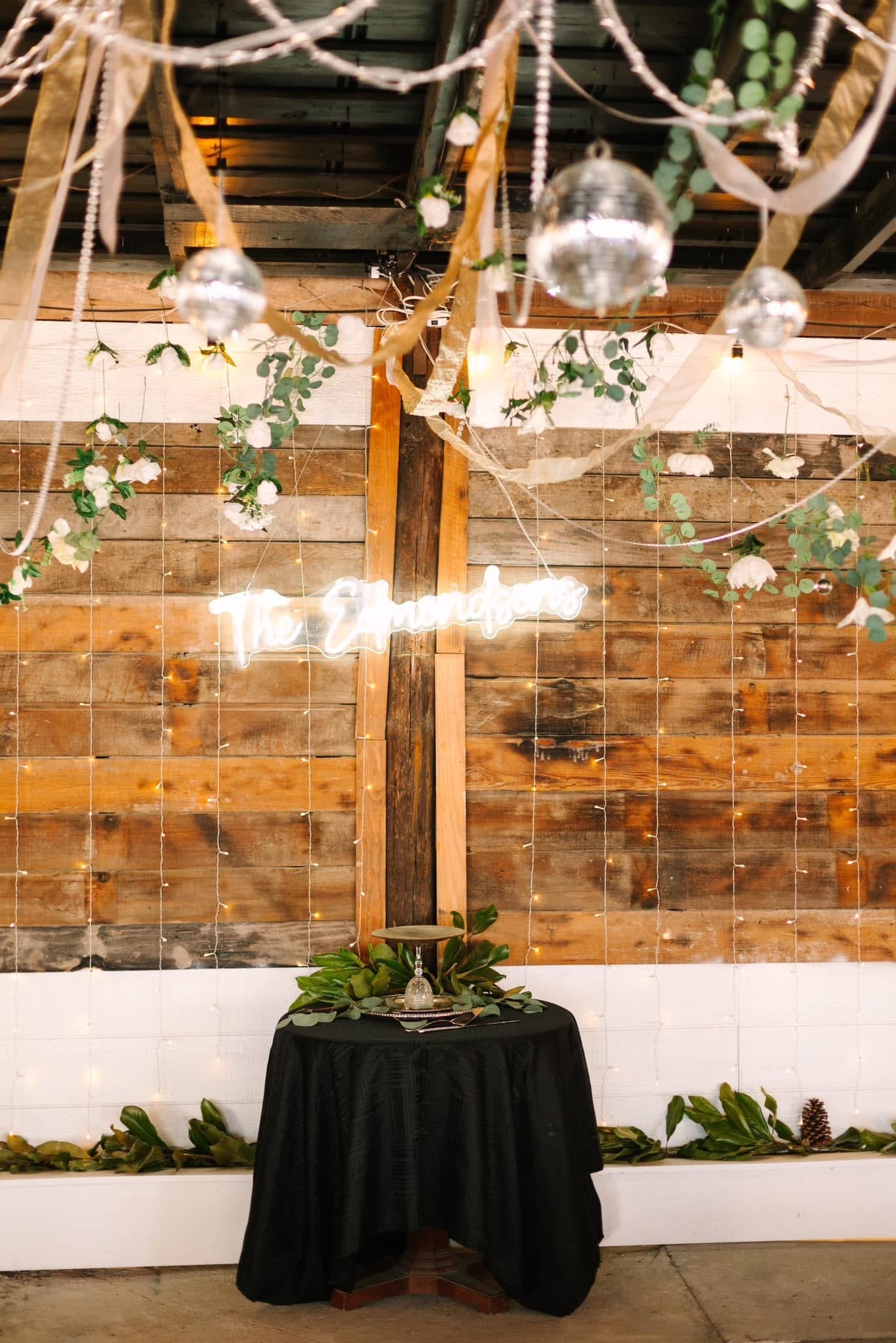 A rustic wedding reception backdrop featuring a glowing neon surname sign on a reclaimed wood wall adorned with fairy lights, white florals, eucalyptus greenery, and hanging disco balls above a black-draped cocktail table.