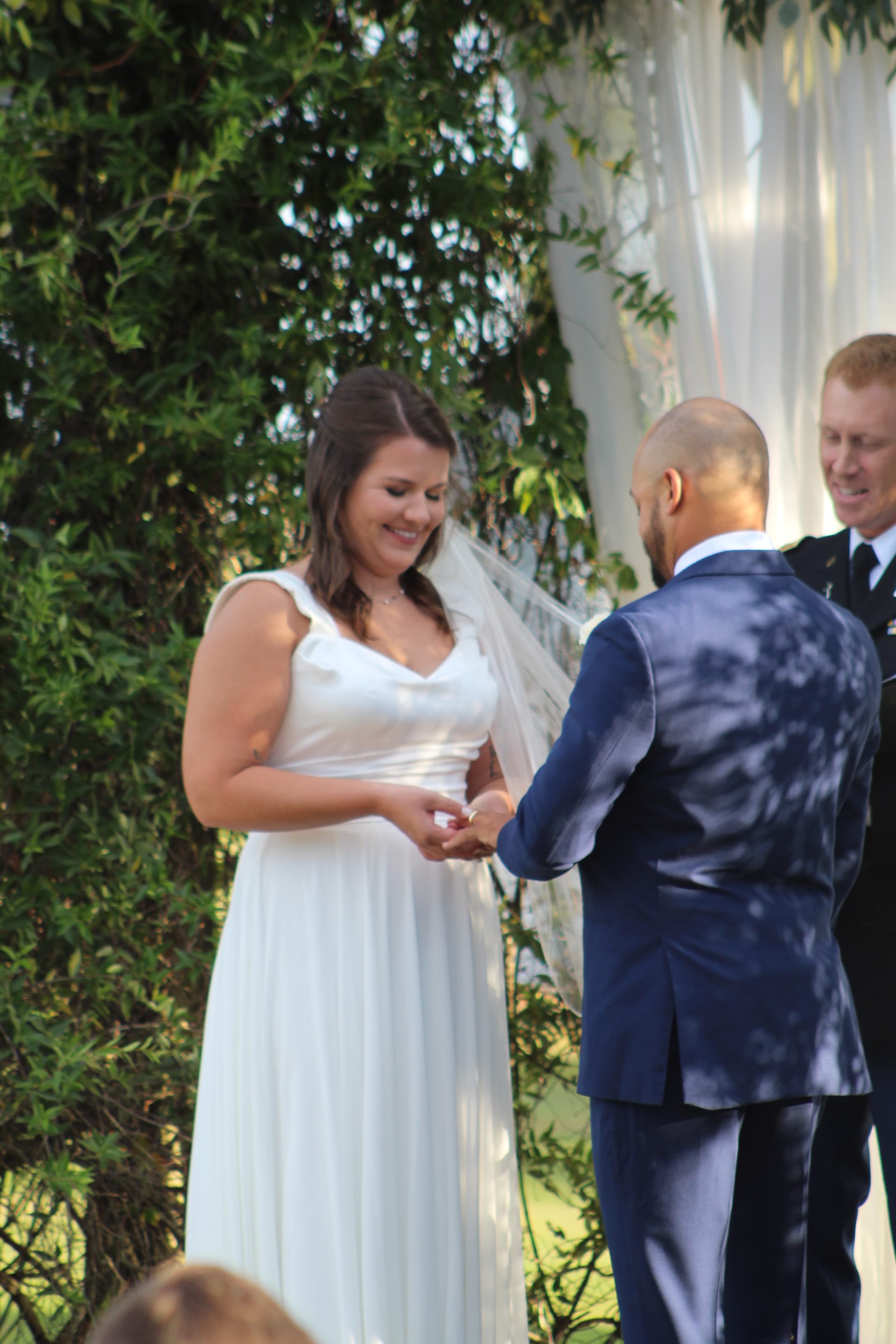 Bride in a flowing white gown and veil smiles as groom places a ring on her finger during an outdoor wedding ceremony at Scarborough House.