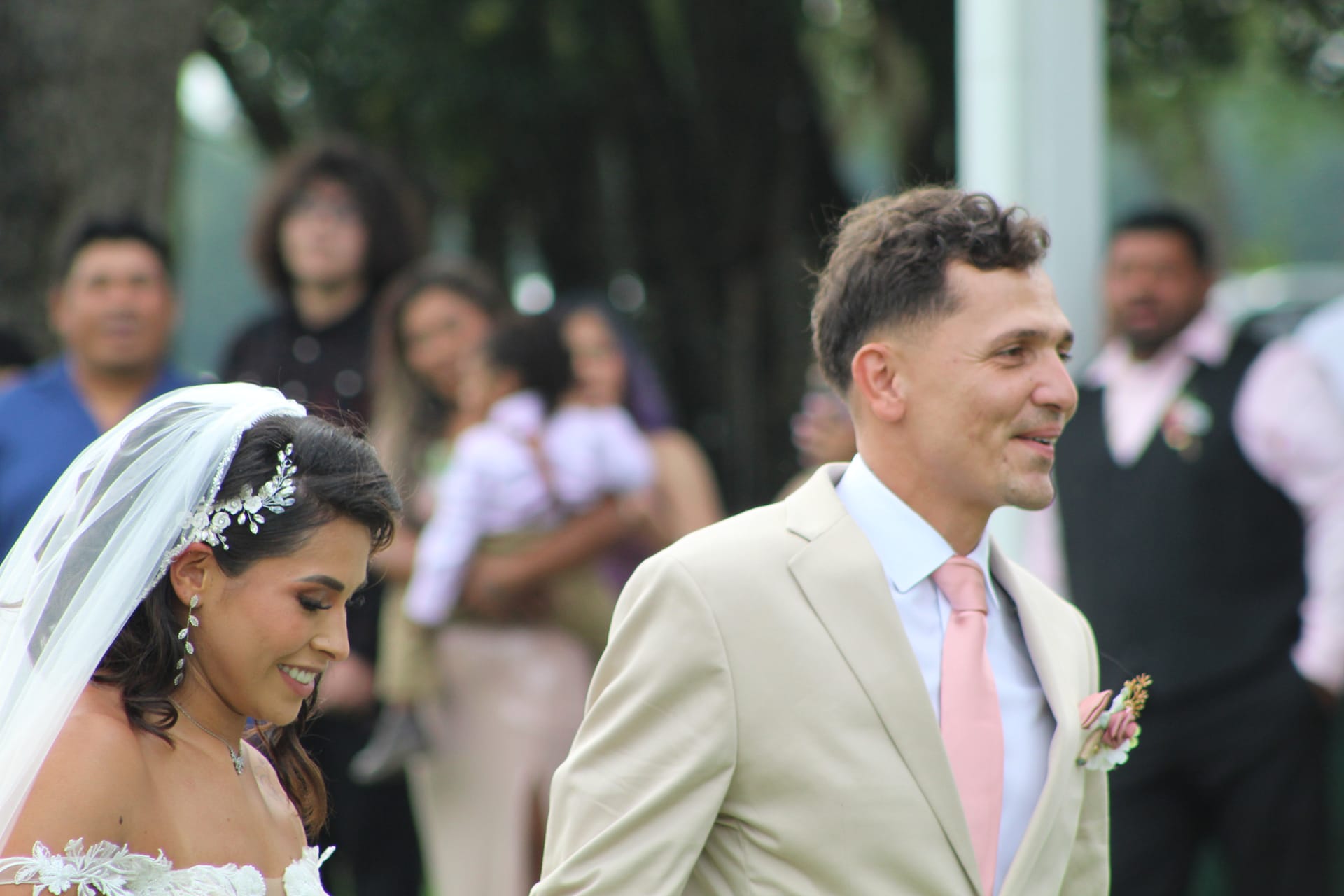 Bride in white lace gown and floral hair piece smiles softly beside groom in tan suit with pink tie during outdoor wedding ceremony at Scarborough House.