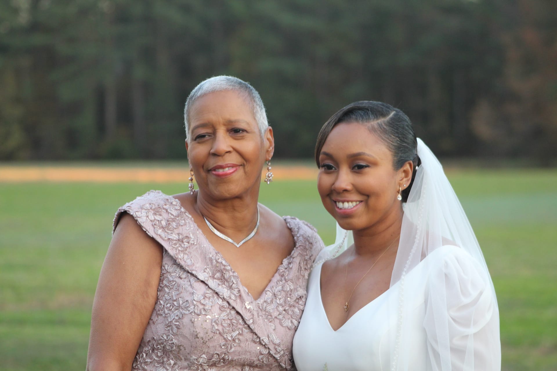 Bride in white gown and veil smiles alongside an older woman in a blush lace dress on the open farmland fields of Scarborough House estate.