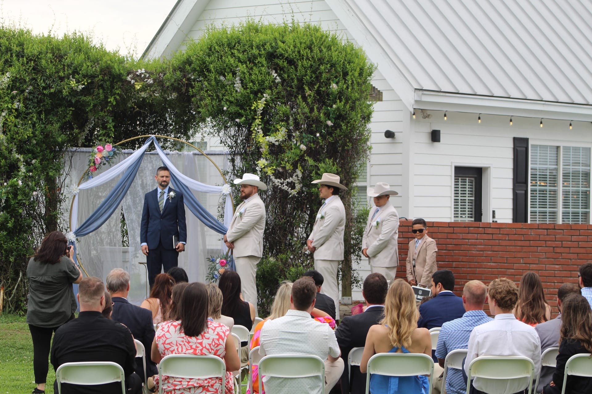 Groom and three groomsmen wearing white cowboy hats stand beneath a floral-draped circular arch on the ceremony lawn at Scarborough House as seated guests look on.